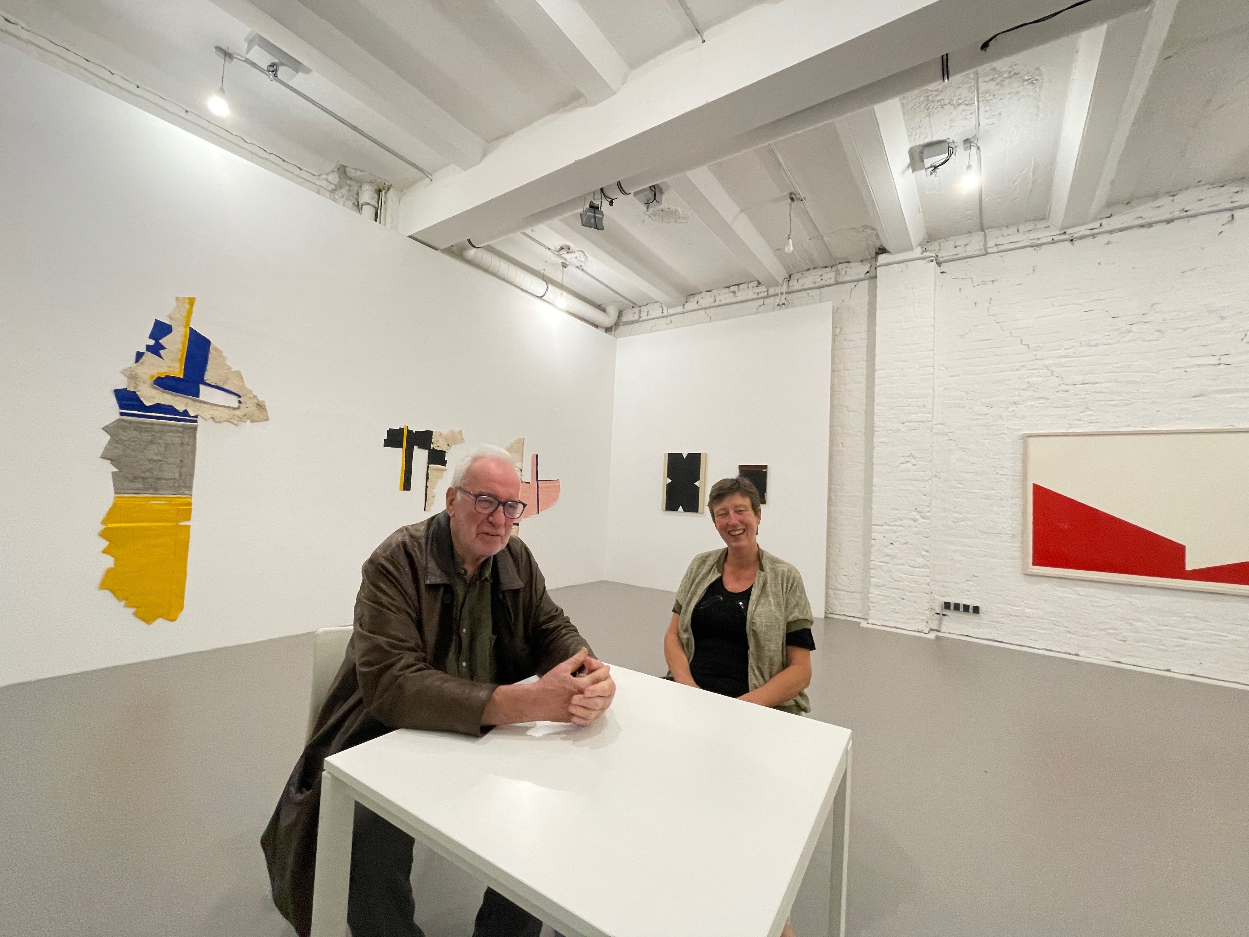 Ein Mann und eine Frau sitzen an einem Tisch in einer Galerie, an den Wänden hängen Kunstwerke.