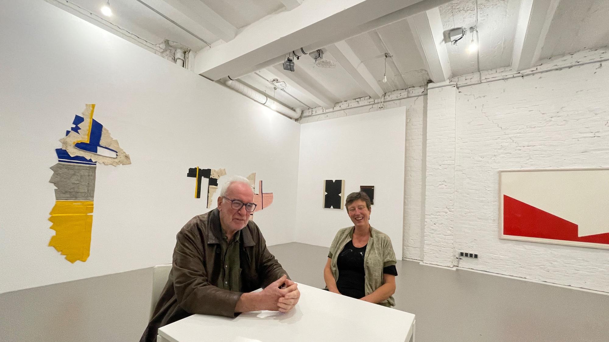 Ein Mann und eine Frau sitzen an einem Tisch in einer Galerie, an den Wänden hängen Kunstwerke.