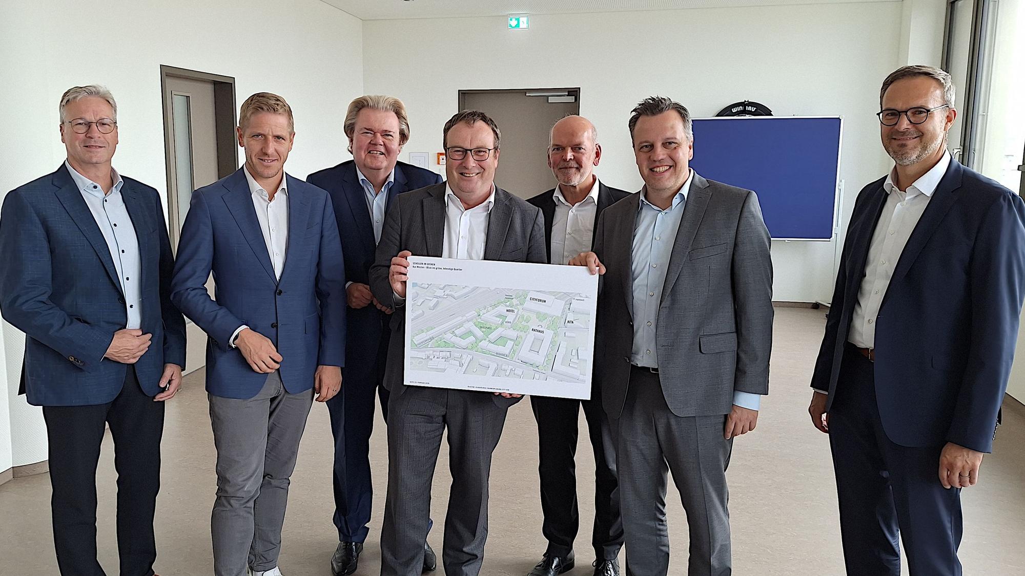 Sieben Männer in Anzügen stehen in einem Raum und zeigen einen Übersichtsplan für das Stadtentwicklungsgebiet "City Süd" in Euskirchen.