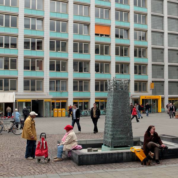 Auf einem Platz steht ein Brunnen, auf der Umrandung sitzen Menschen.