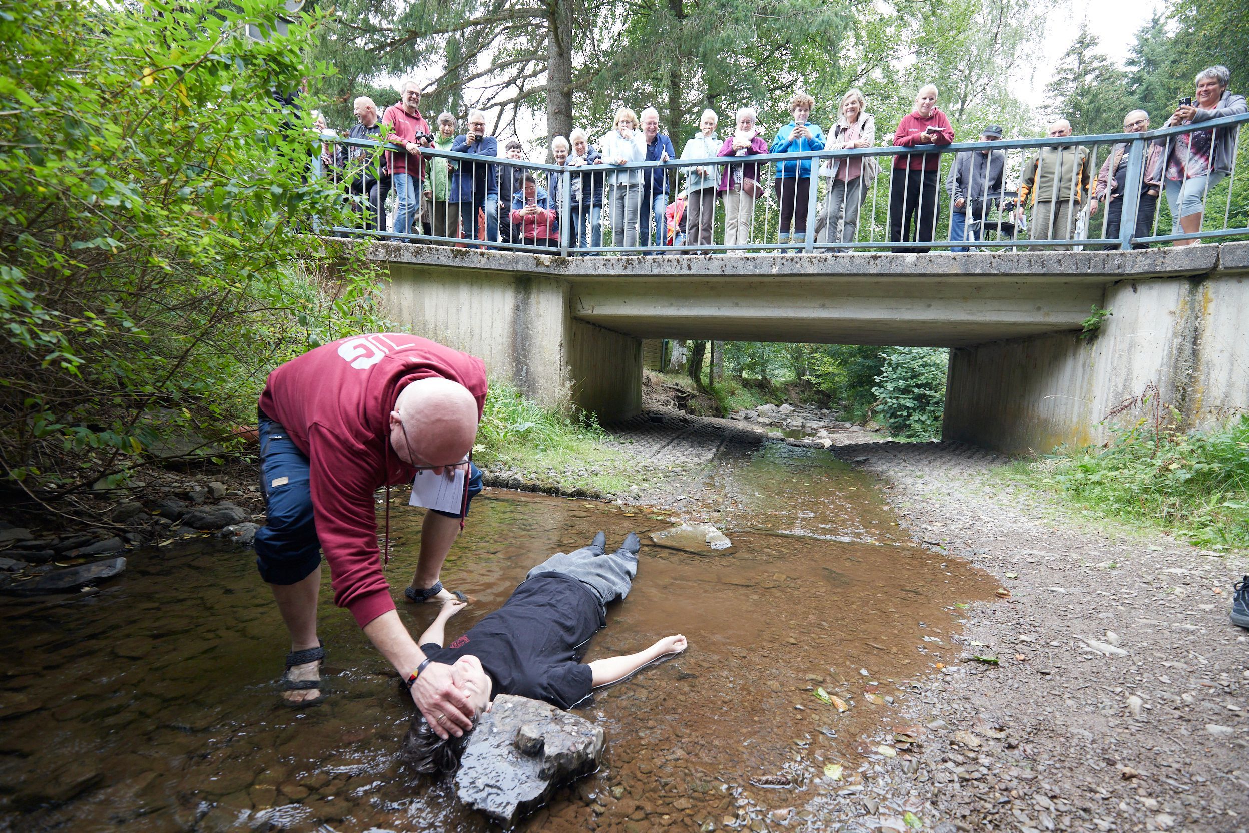 Ein Jugendlicher liegt in einem Bach, der sehr wenig Wasser führt, und wird vom Pfarrer getauft.