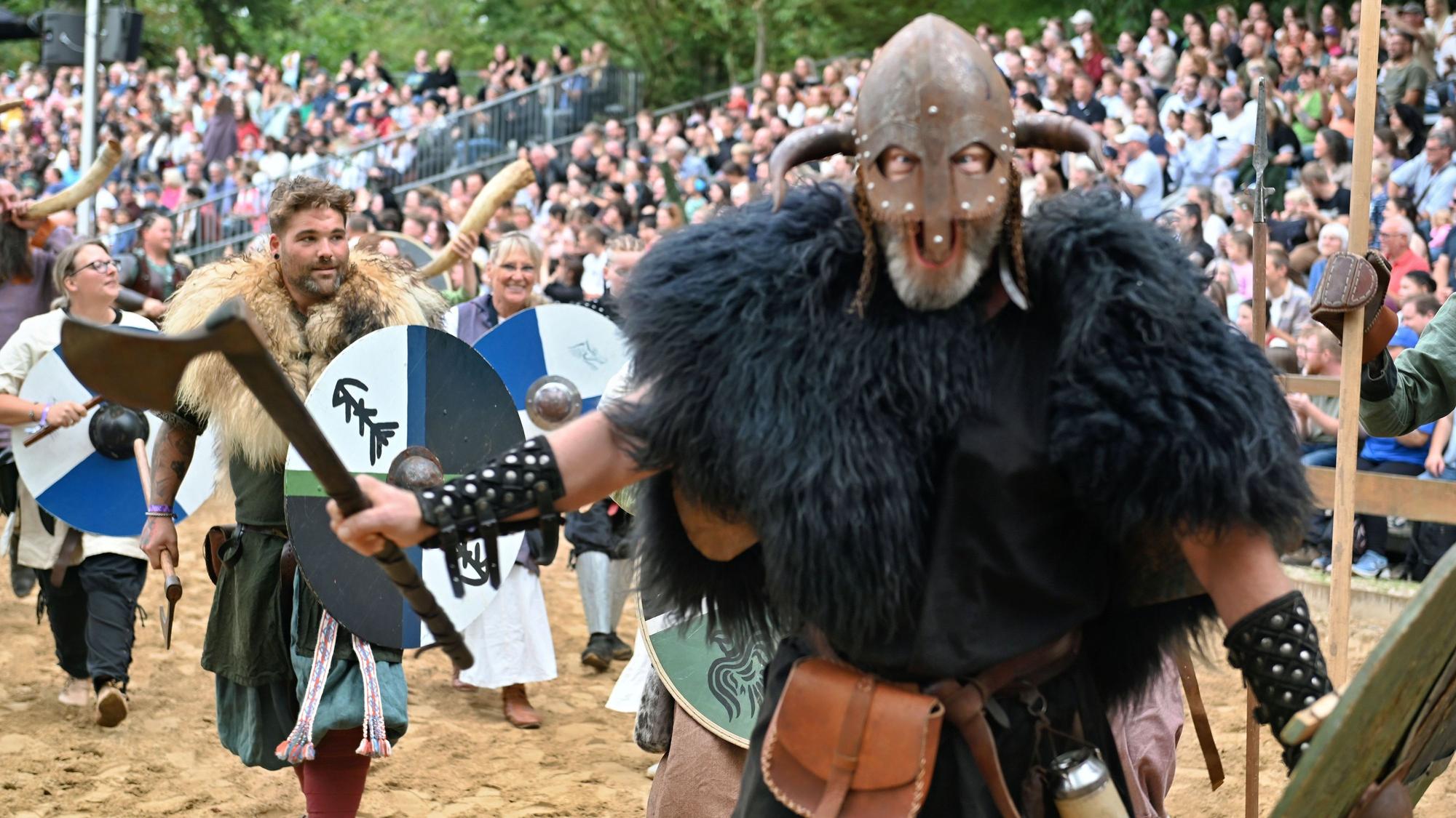 Ein Mann in schwarzem Fell und mit einer Maske mit Hörnern läuft gröhlend mit einer Axt in der Hand auf die Kamera zu, im Hintergrund sieht man weitere mittelalterlich gekleidete Leute.