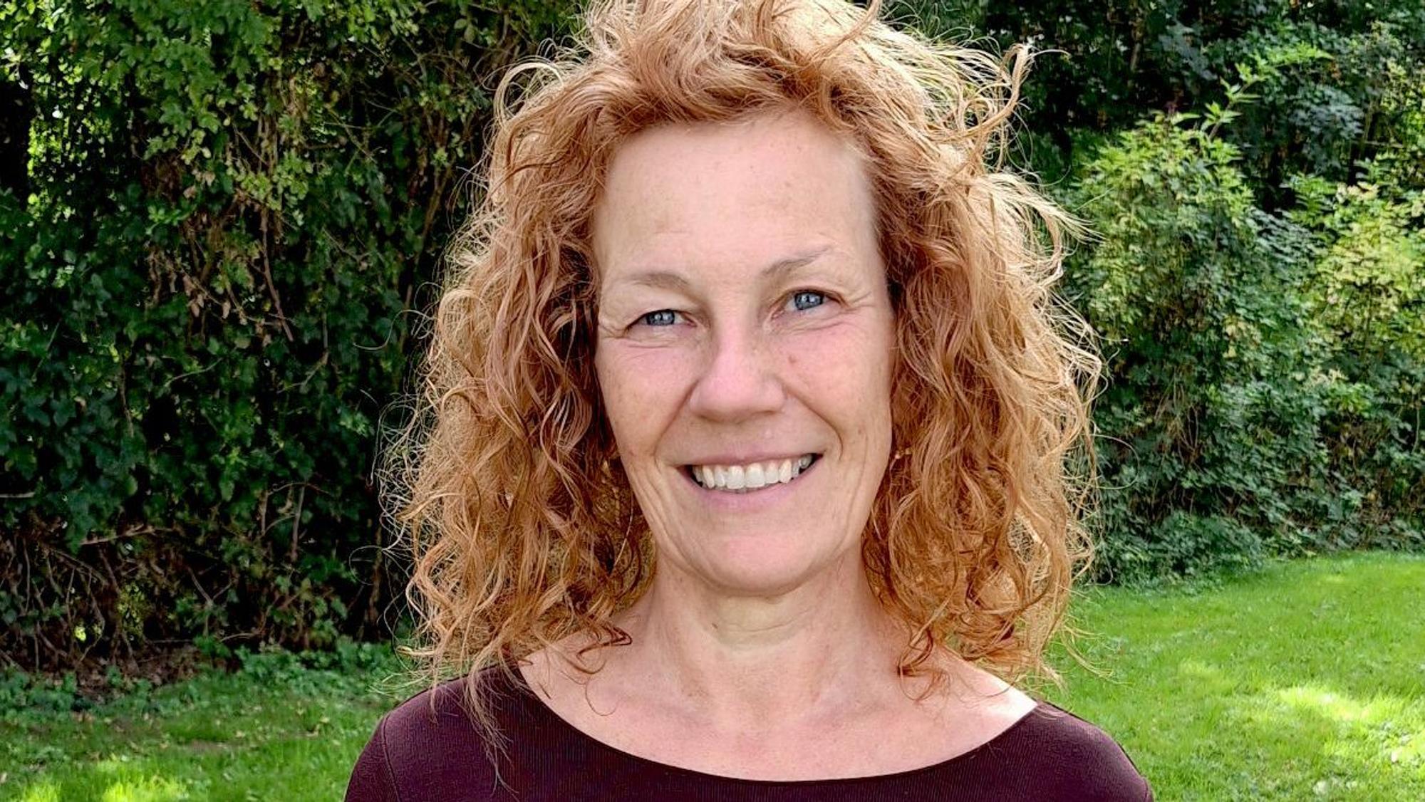Die Frau in einem dunkelroten Oberteil und roten Locken steht auf einer Wiese.