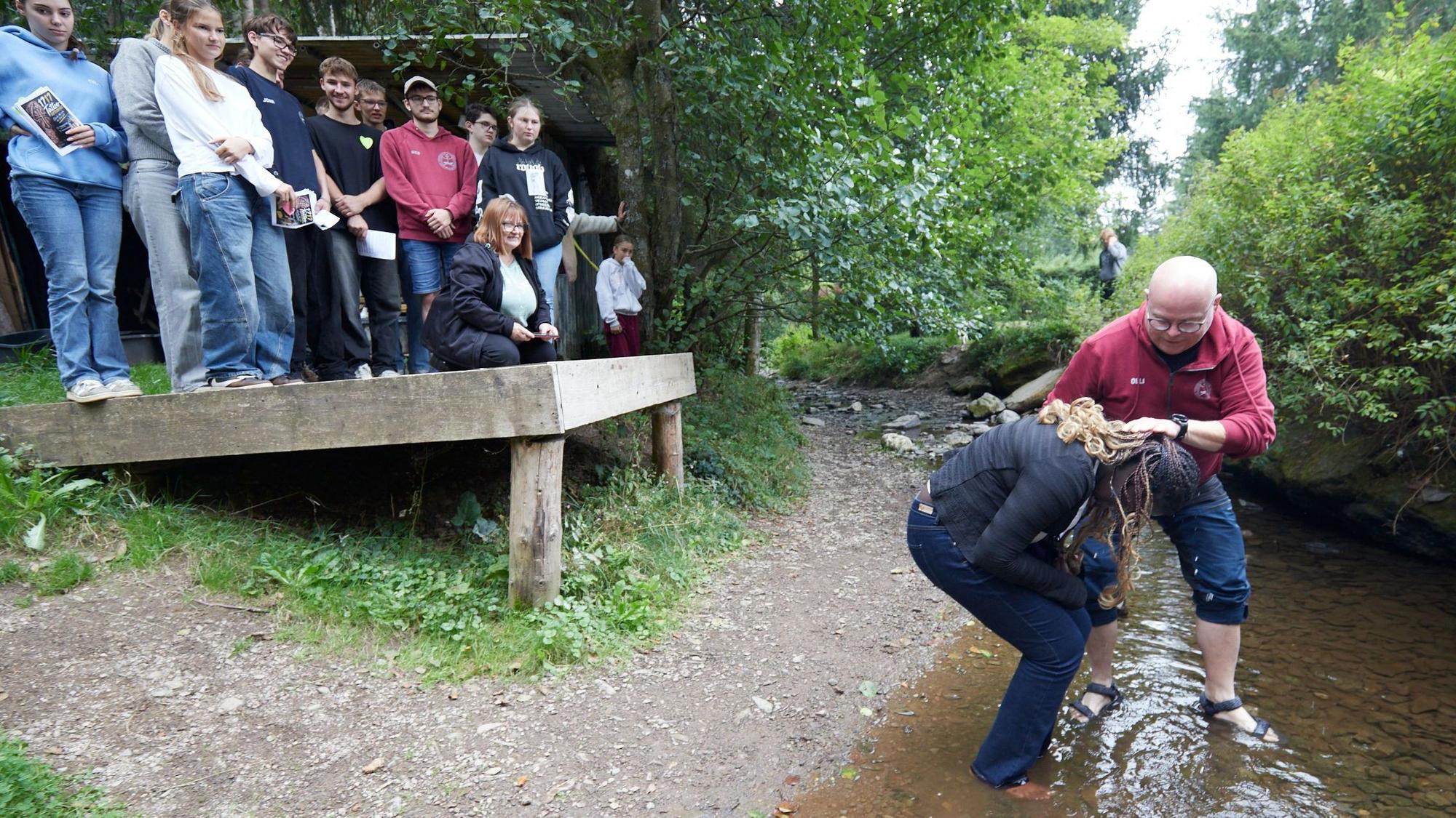 Die junge Frau steht in einem Bach und wird vom Pfarrer getauft, derr Wasser über ihren Kopf schüttet.