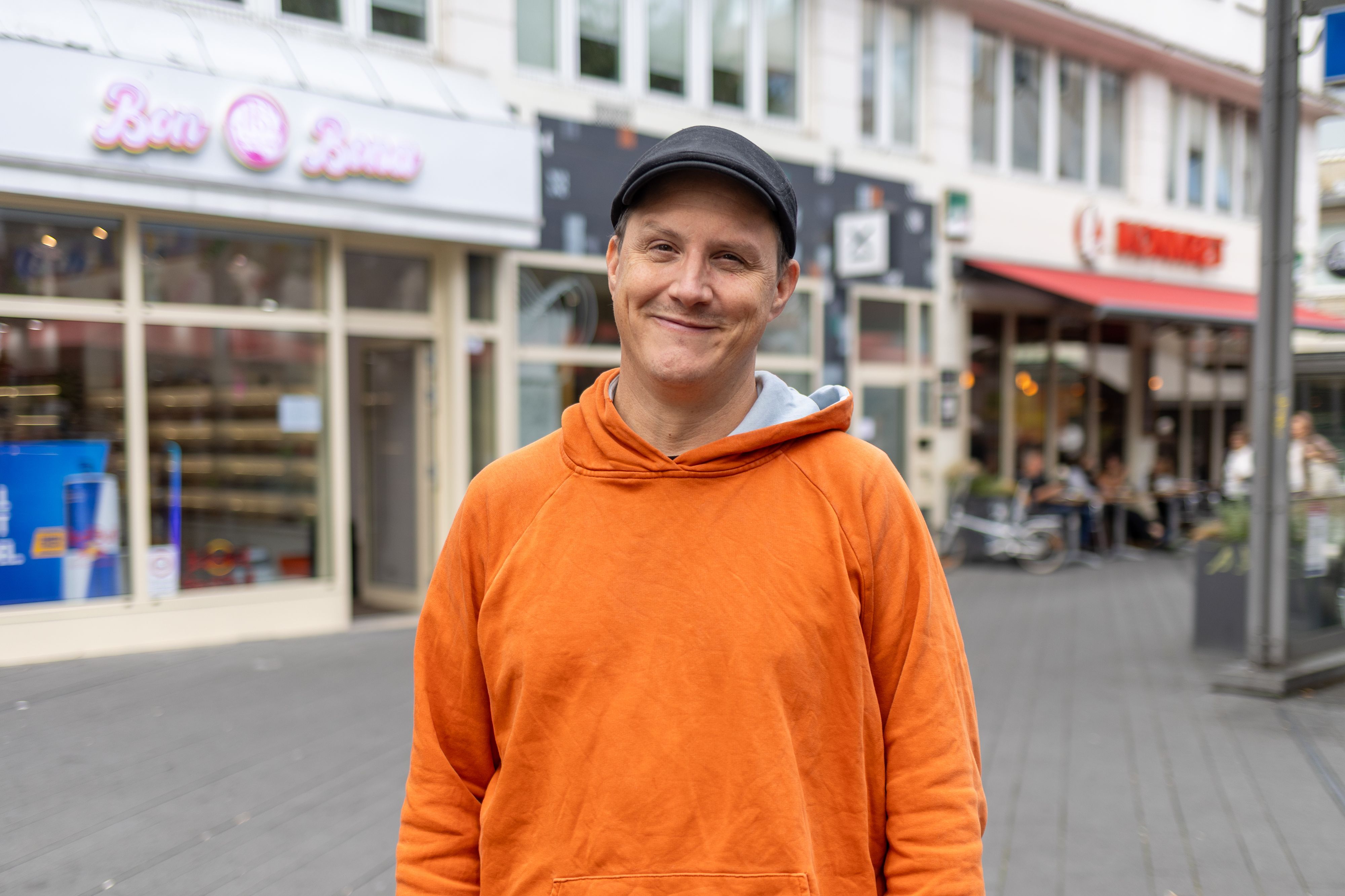 Ein Mann im orangefarbenen Pullover lächelt in die Kamera.