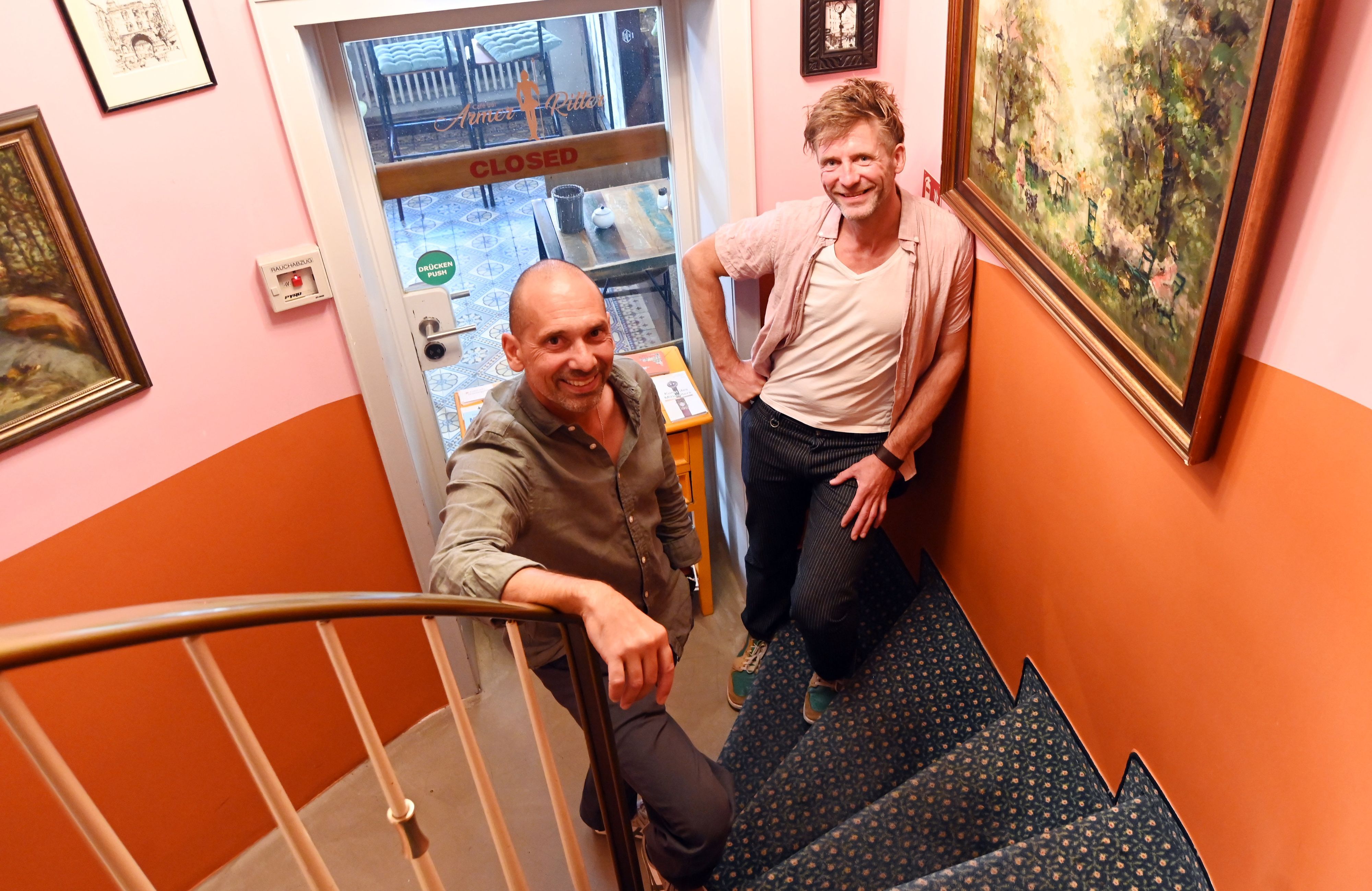 Zwei Männer stehen auf der mit Teppich belegten Treppe eines Hotels. An den Wänden hängen altertümlich anmutende Bilder.