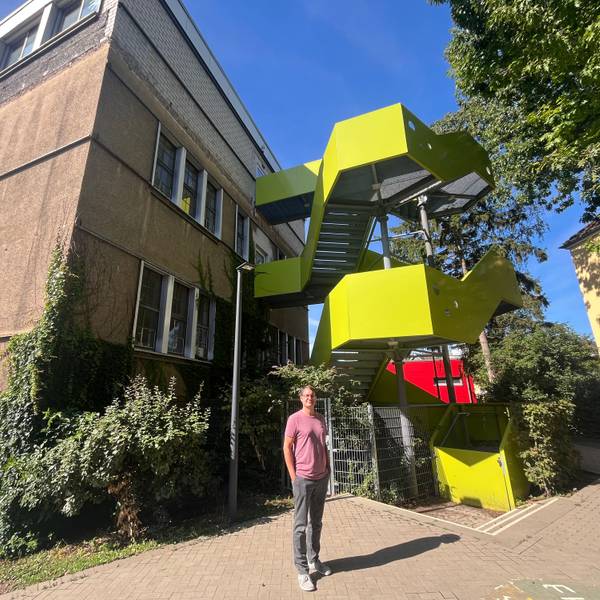 Ein Mann steht vor einem Schulgebäude mit einer rotgelben Feuertreppe.