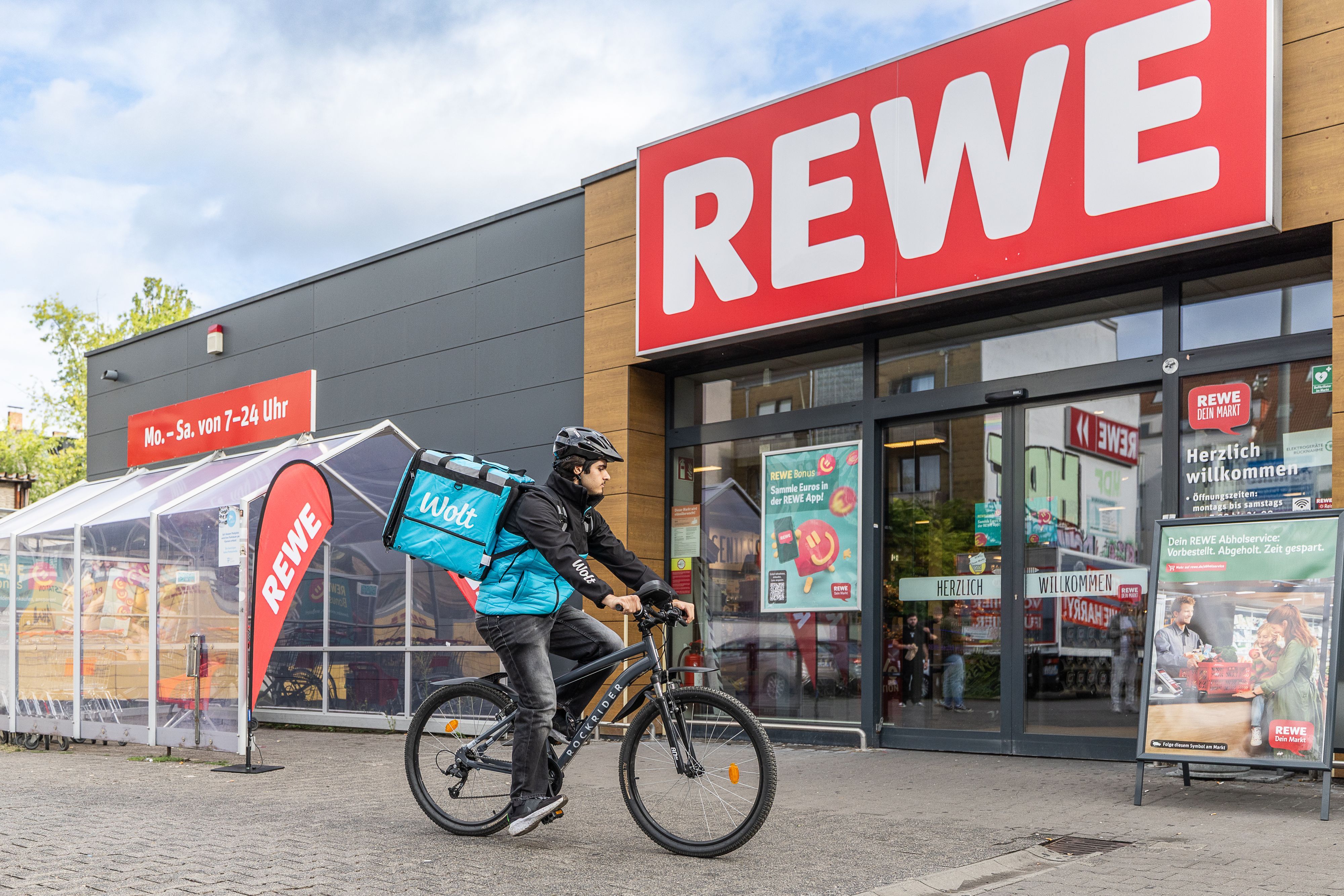 Ein Wolt-Fahrer hält auf seinem Rad vor einer Rewe-Filiale.