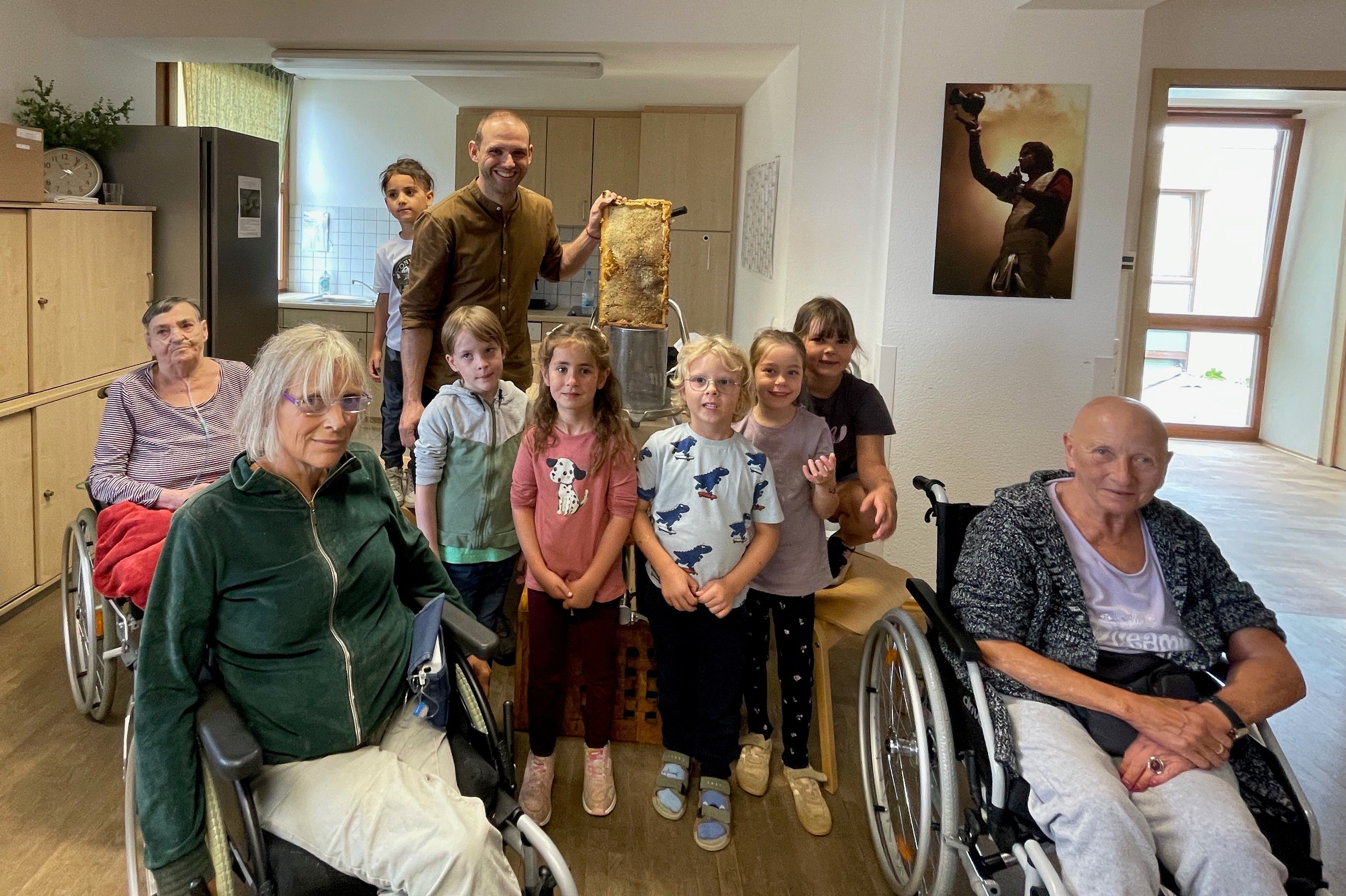 Fünf Kinder der Kita „Alexianer Pänz“ stehen mit zwei Hospiz-Bewohnerinnen, die im Rollstuhl sitzen, in der Küche des Alexianer-Hospiz in Köln-Rondorf