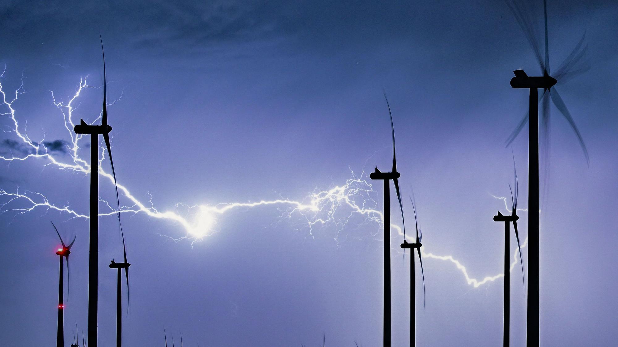 Das Symbolfoto zeigt einen mächtigen verzweigten Gewitterblitz, der die nächtliche Landschaft eines Windparks erleuchtet.