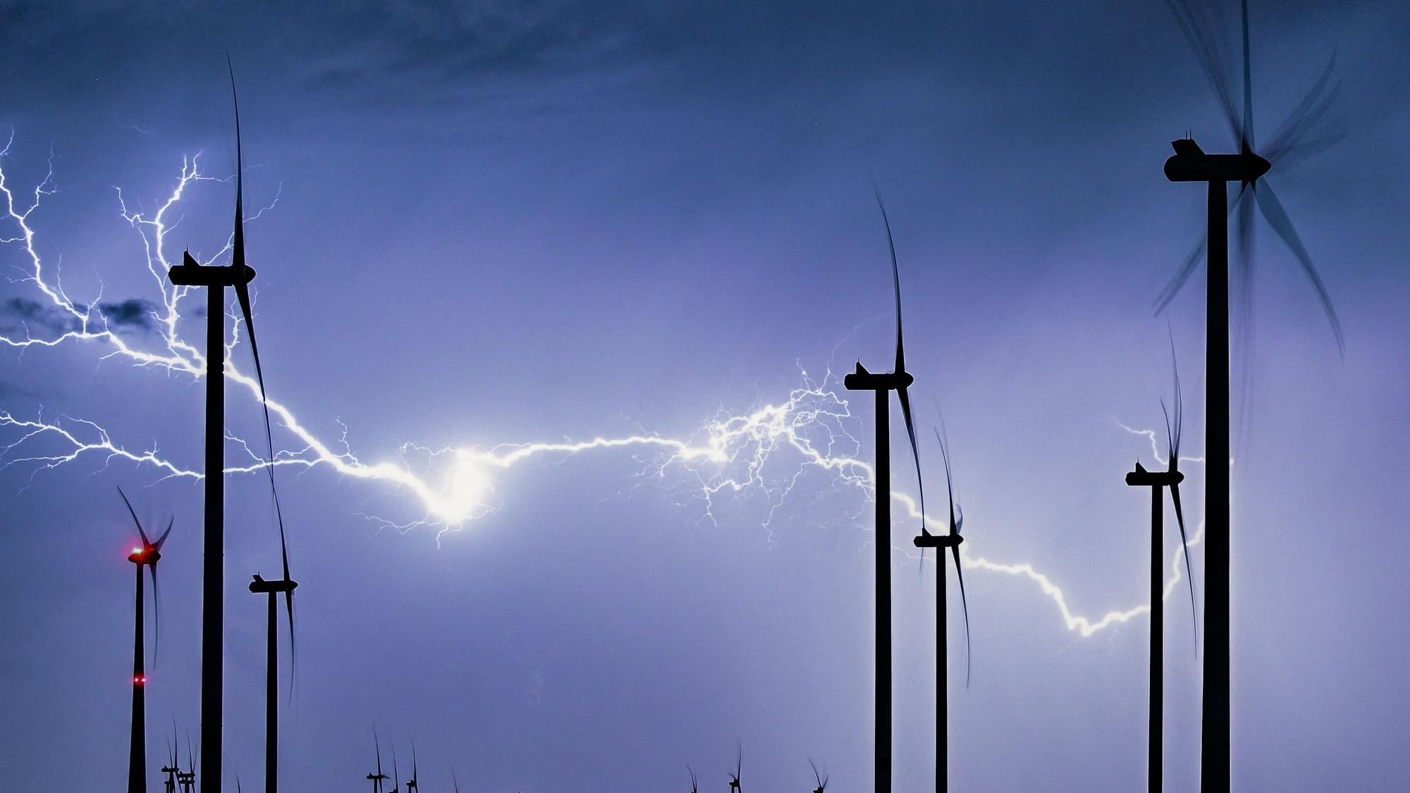 Ein mächtiger, verzweigter Gewitterblitz leuchtet über der nächtlichen Landschaft mit einem Windpark.