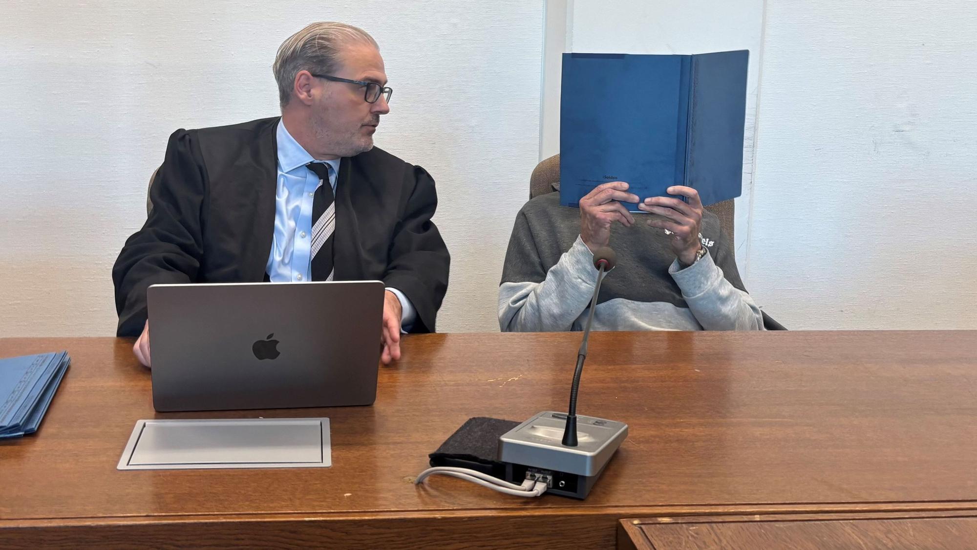 Zu sehen sind der Verteidiger und der Angeklagte, der sich eine Kladde vor sein Gesicht hält.