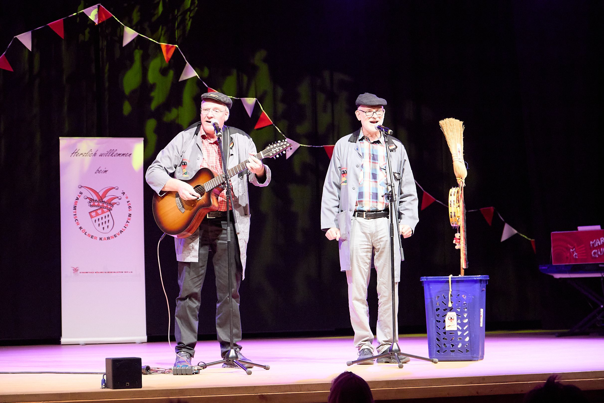 Zwei Männer stehen auf einer Bühne und sprechen in Mikrofone. Einer spielt Gitarre.