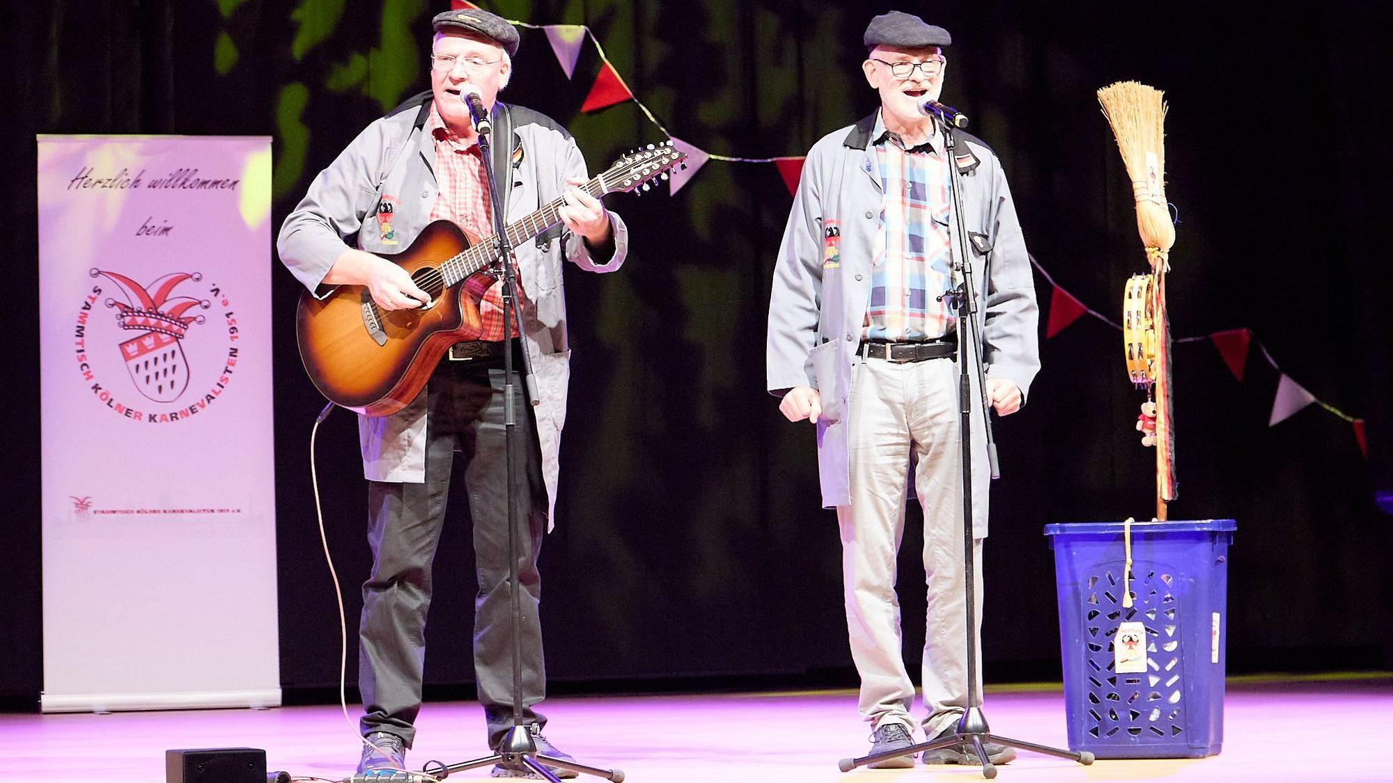 Zwei Männer stehen auf einer Bühne und sprechen in Mikrofone. Einer spielt Gitarre.