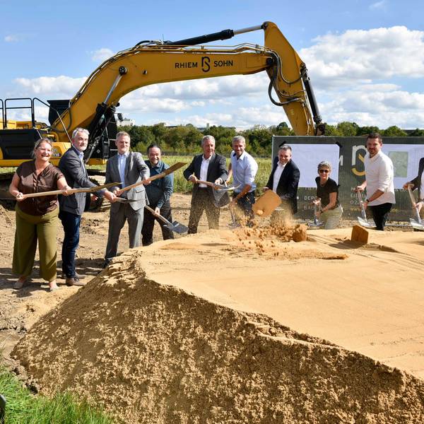 Das Foto zeigt den symbolischen ersten Spatenstich mit Bagger im Hintergrund.