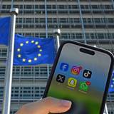 Eine Person hält ein Smartphone mit den Logos der Social-Media-Apps Facebook, Instagram, TikTok, X, Snapchat und WhatsApp vor Fahnen der Europäischen Union und dem Gebäude der Europäischen Kommission. Die EU-Kommission will den Zugang zu Social Media für Kinder einschränken.