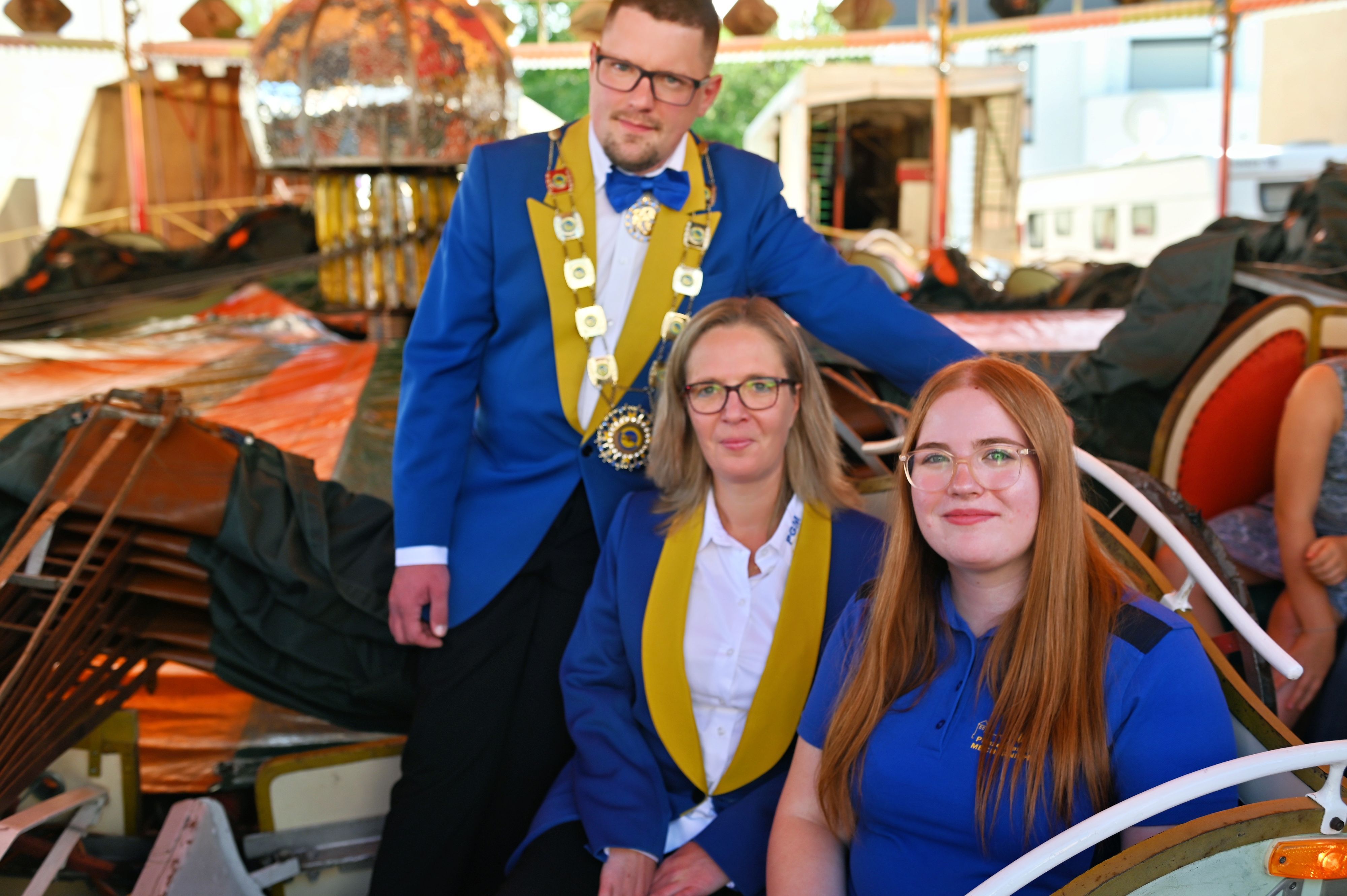 Die drei Genannten tragen das blaue Outfit ihres Karnevalsvereins und posieren vor einem Kirmeskarussell für den Fotografen.