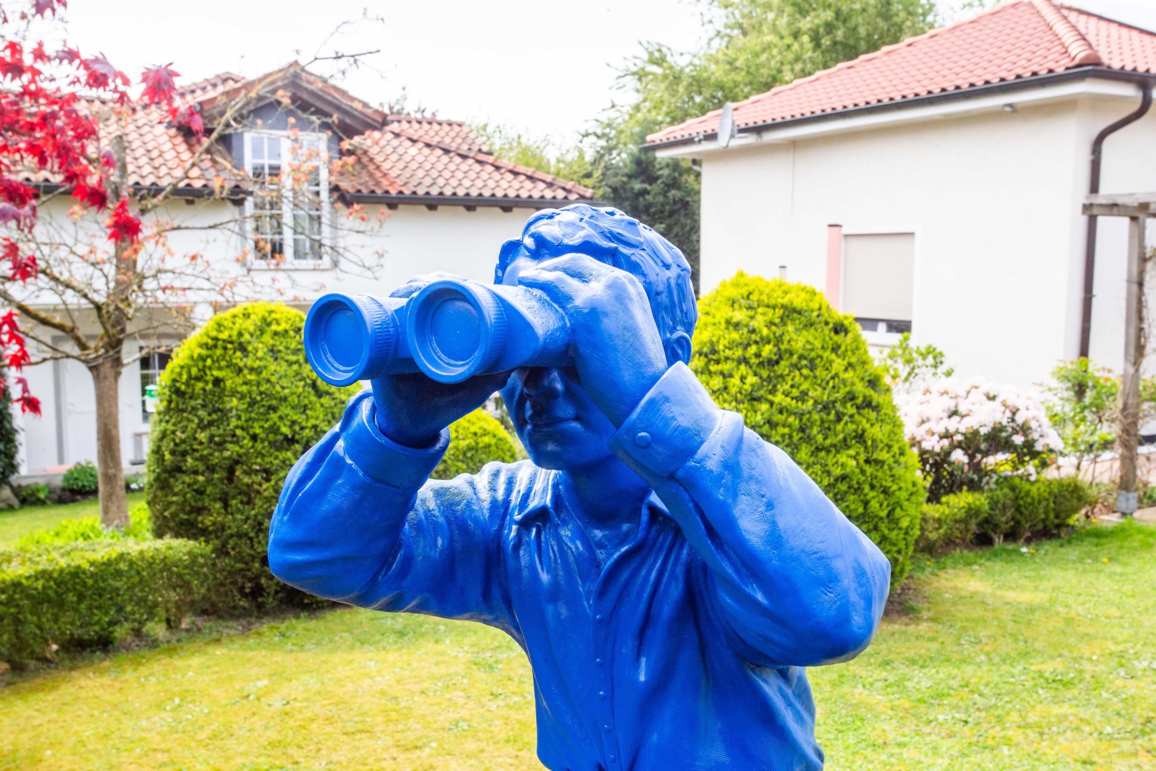 Eine blaue Statue eines Mannes, der durch ein Fernglas schaut, ist in einem Garten zu sehen.
