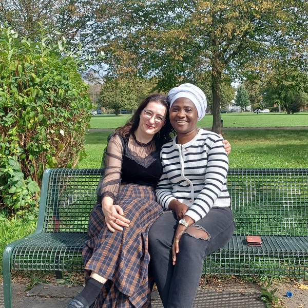 Eine junge Frau mit dunklem Haar und langem Rock sitzt neben einer Schwarzen Frau mit Turban und Jeans auf einer Parkbank.