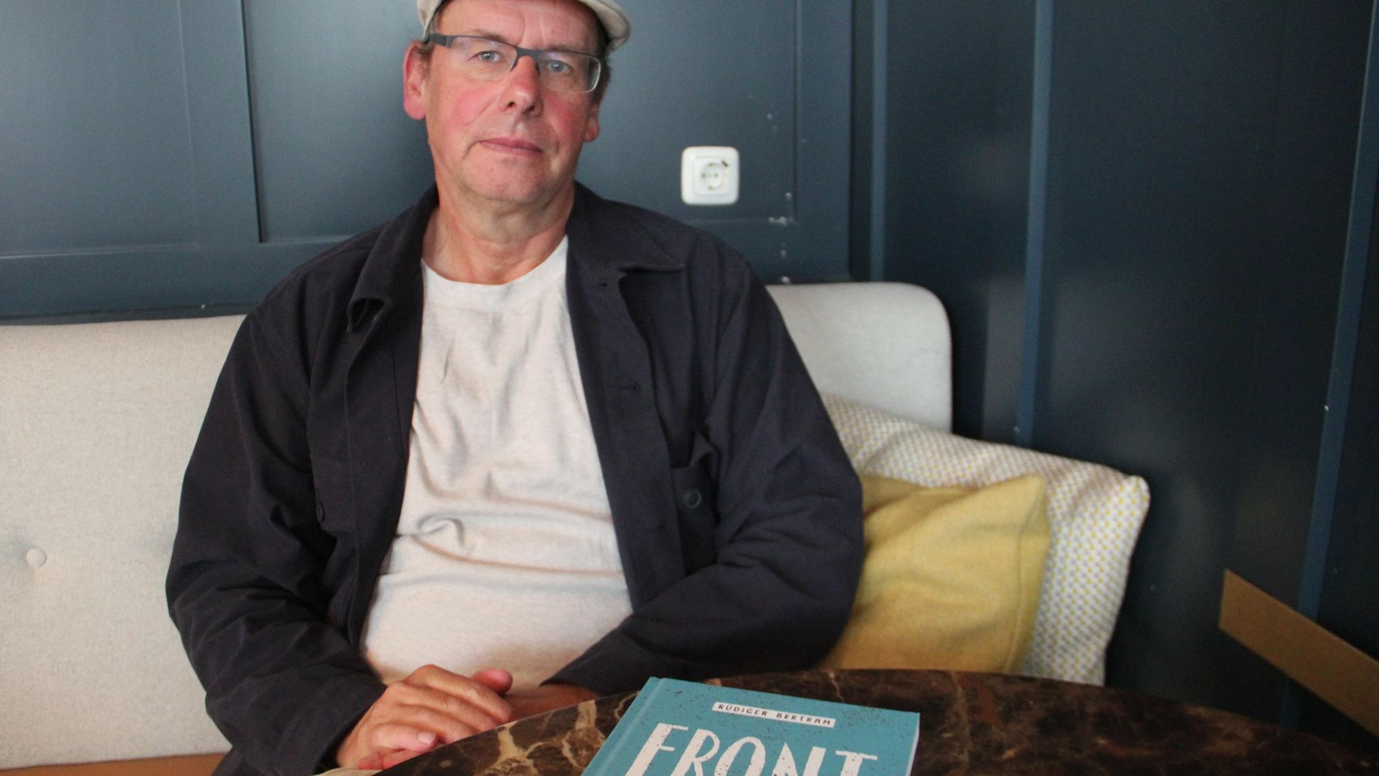 Ein Mann mit Kappe und Brille sitzt auf einer Bank. Vor ihm auf dem Tisch liegt sein neues Buch „Front“.
