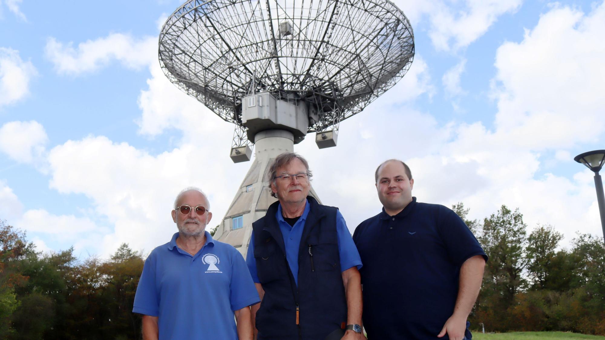 Drei Männer stehen nebeneinander, im Hintergrund thront der 25-Meter-Spiegel des Astropeilers.