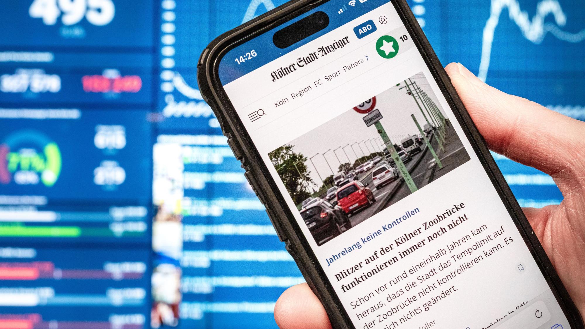 13.02.2025, Köln: Die Webseite des Kölner Stadt-Anzeigers ksta.de ist auf einem Smartphone zu sehen. Im Hintergrund ist ein Monitor im Newsroom der Tageszeitung sichtbar. Foto: Matthias Heinekamp