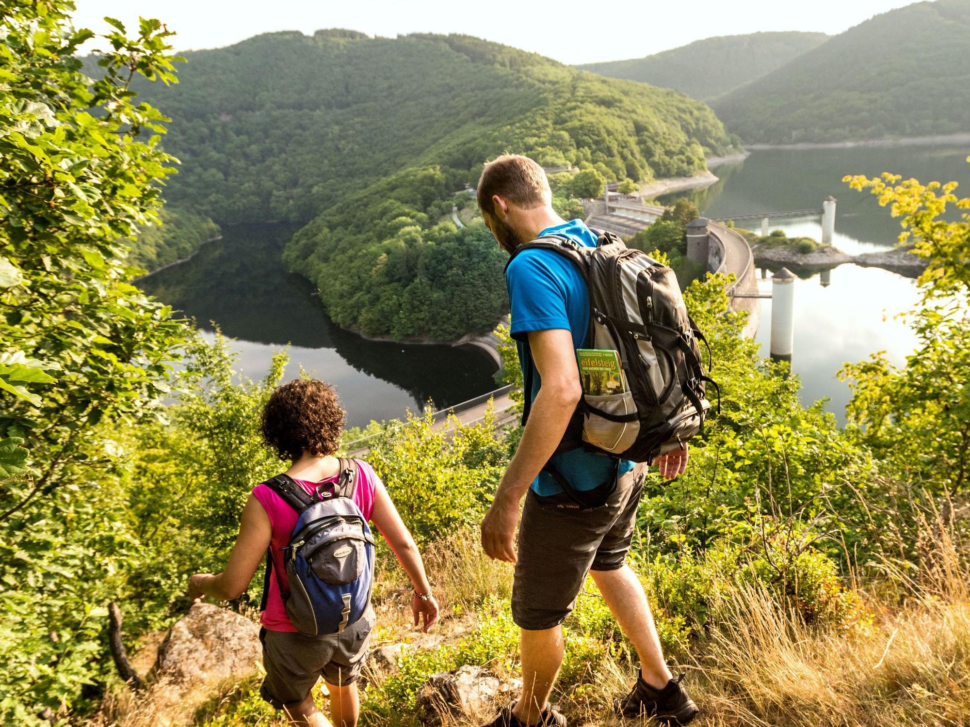 Zwei Wanderer mit Rucksäcken, die auf dem Foto nur mit dem Rücken zu sehen sind, steigen einen Weg in Richtung der Staumauer des Urftsees hinab.