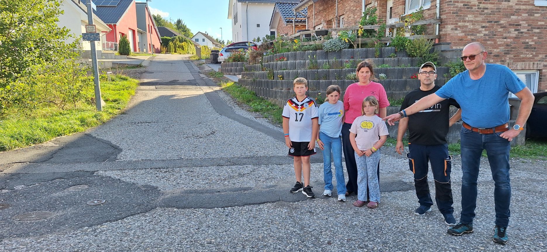 Die genannten Personen stehen an einer Erschließungsstraße in einem Neubaugebiet, Matthias Stegemann deutet auf Schäden der groben Asphaltdecke.