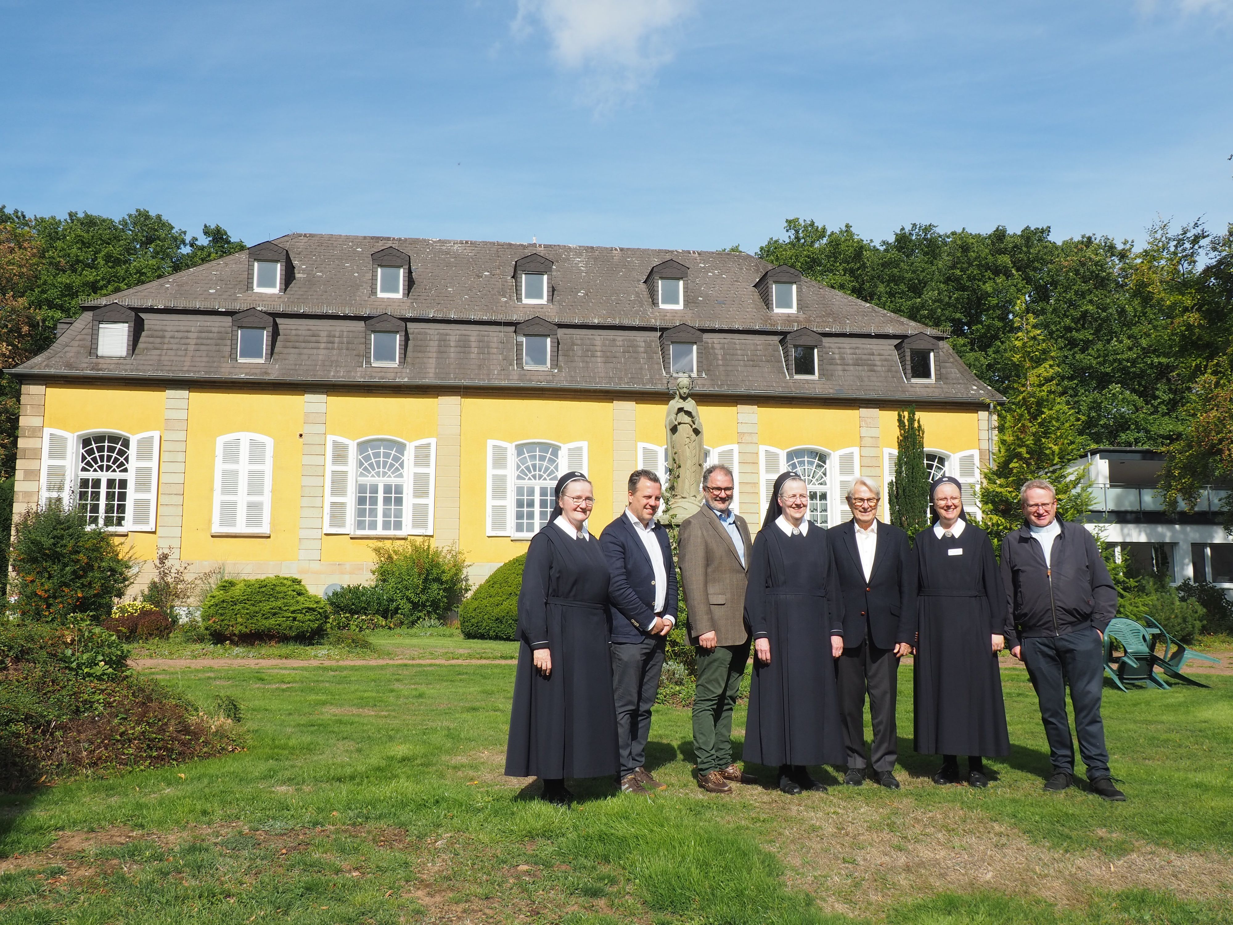 Eine Gruppe Menschen steht auf einer Wiese vor dem Haupthaus vonm Maria Wald.