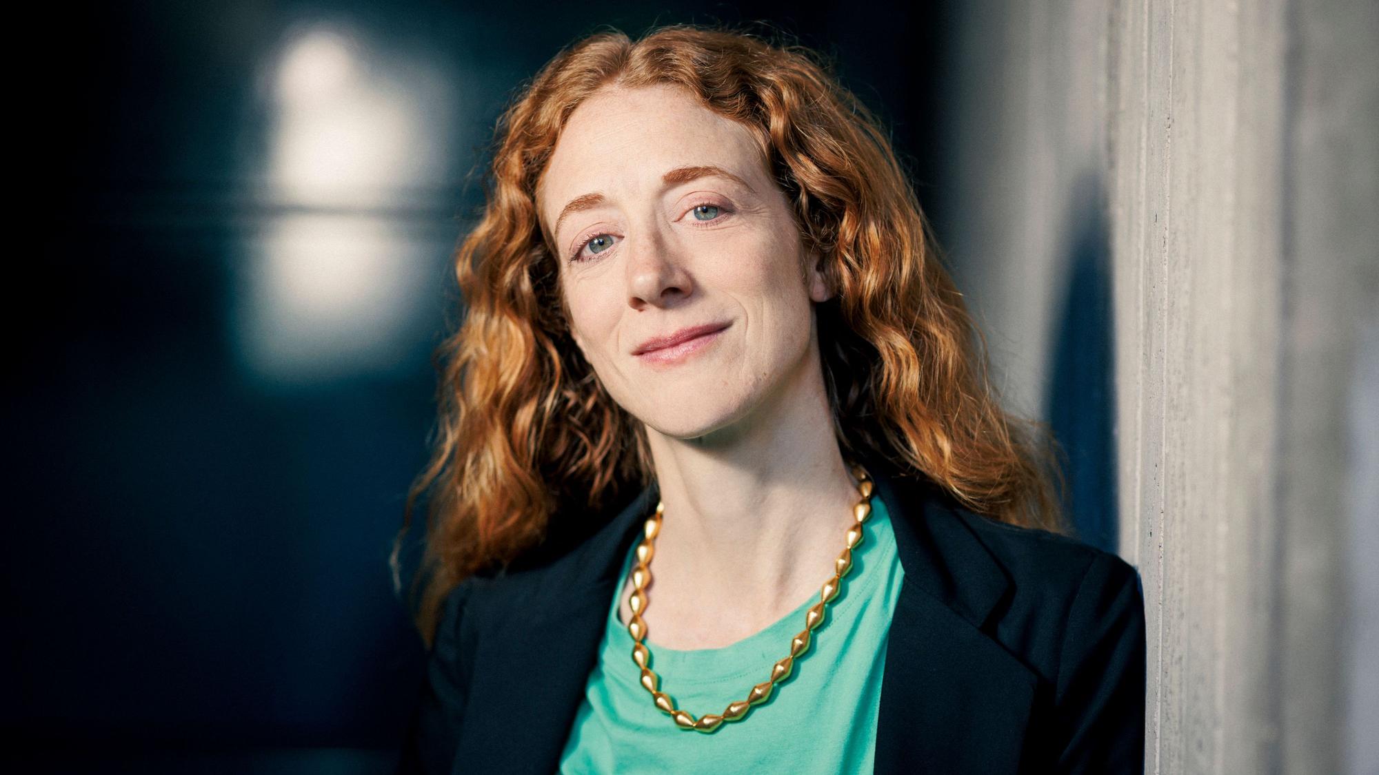 Lily Sykes im Porträt. Sie lächelt, trägt einen dunklen Blazer und eine grüne Bluse, die mit ihren roten Haaren kontrastiert.