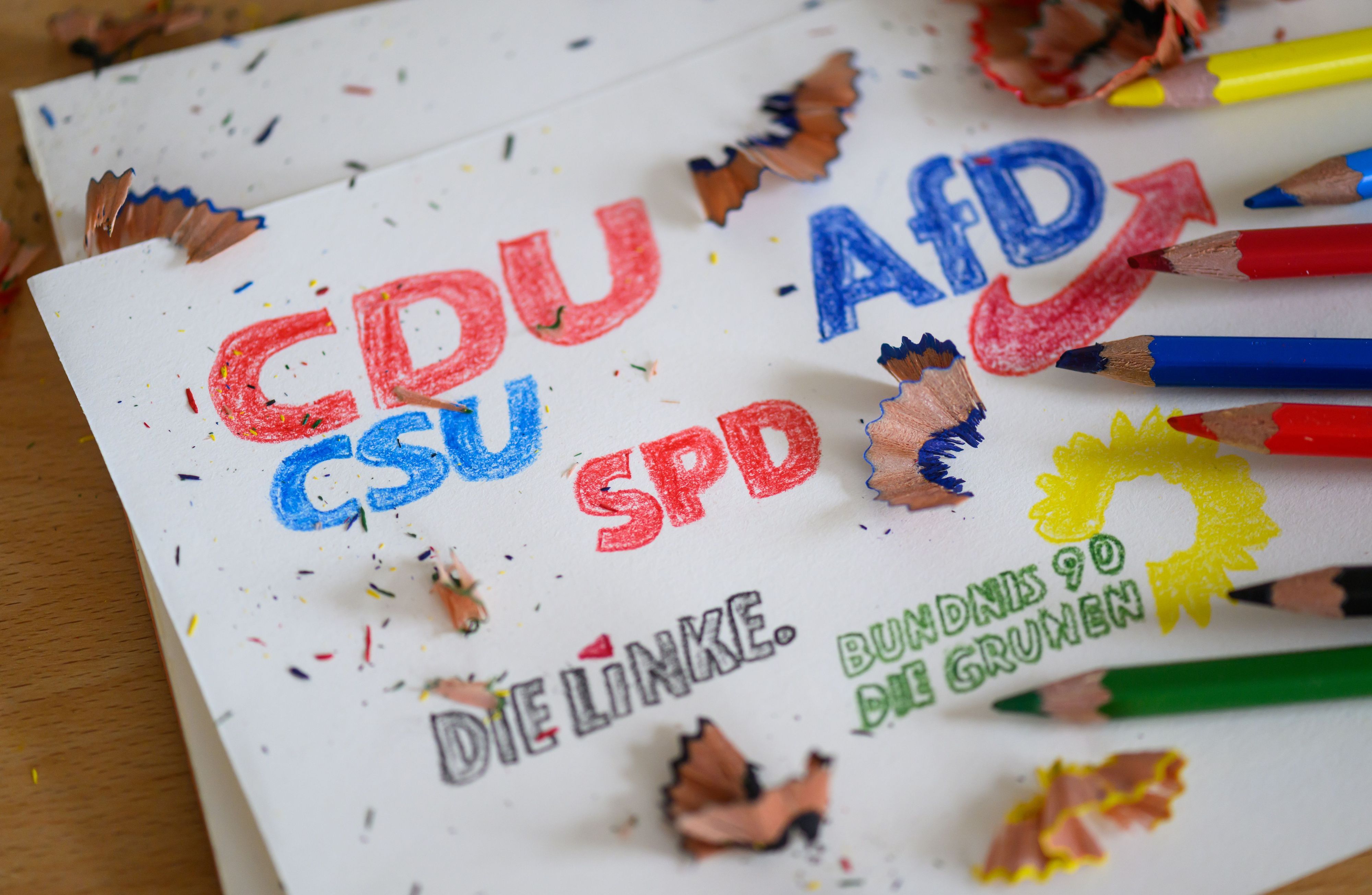 Die Logos der Parteien CDU/CSU, SPD, Die Linke, Bündnis 90/Die Grünen und AfD sind mit Buntstiften auf ein Blatt gezeichnet.