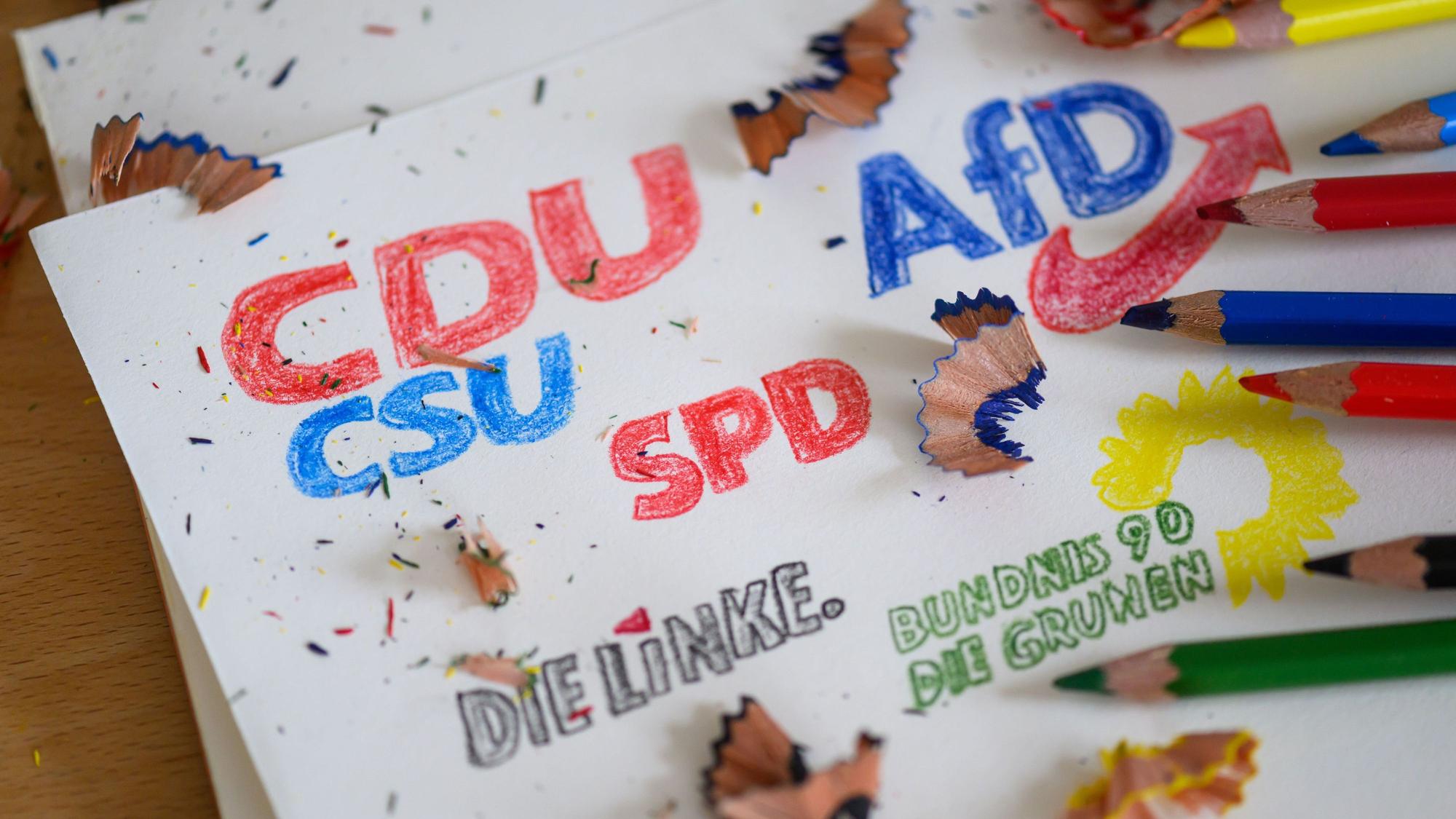 Die Logos der Parteien CDU/CSU, SPD, Die Linke, Bündnis 90/Die Grünen und AfD sind mit Buntstiften auf ein Blatt gezeichnet.