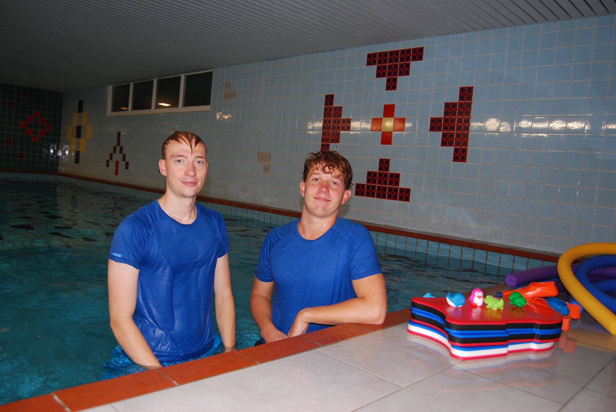 Zwei junge Männer in nassen T-Shirts stehen bis zum Bauch in einem Schwimmbecken.