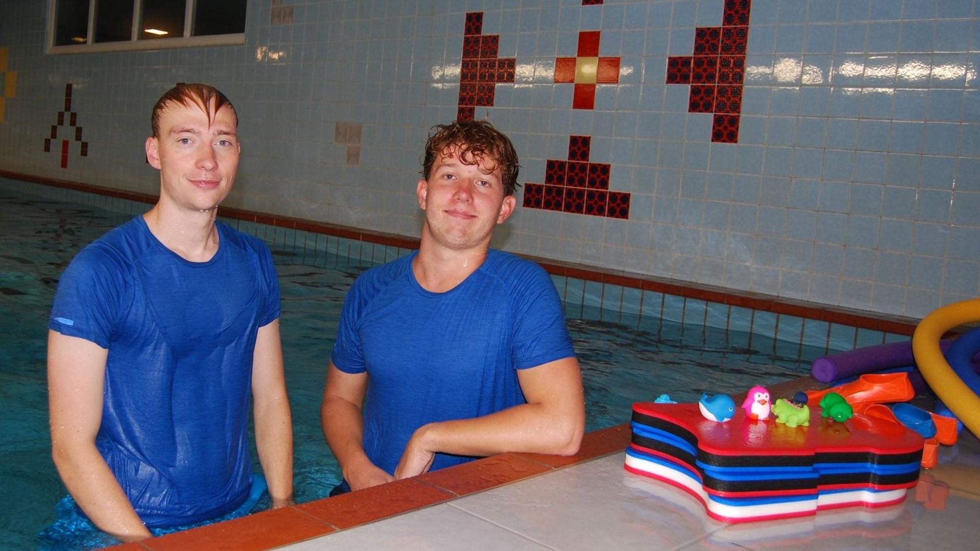 Zwei junge Männer in nassen T-Shirts stehen bis zum Bauch in einem Schwimmbecken.