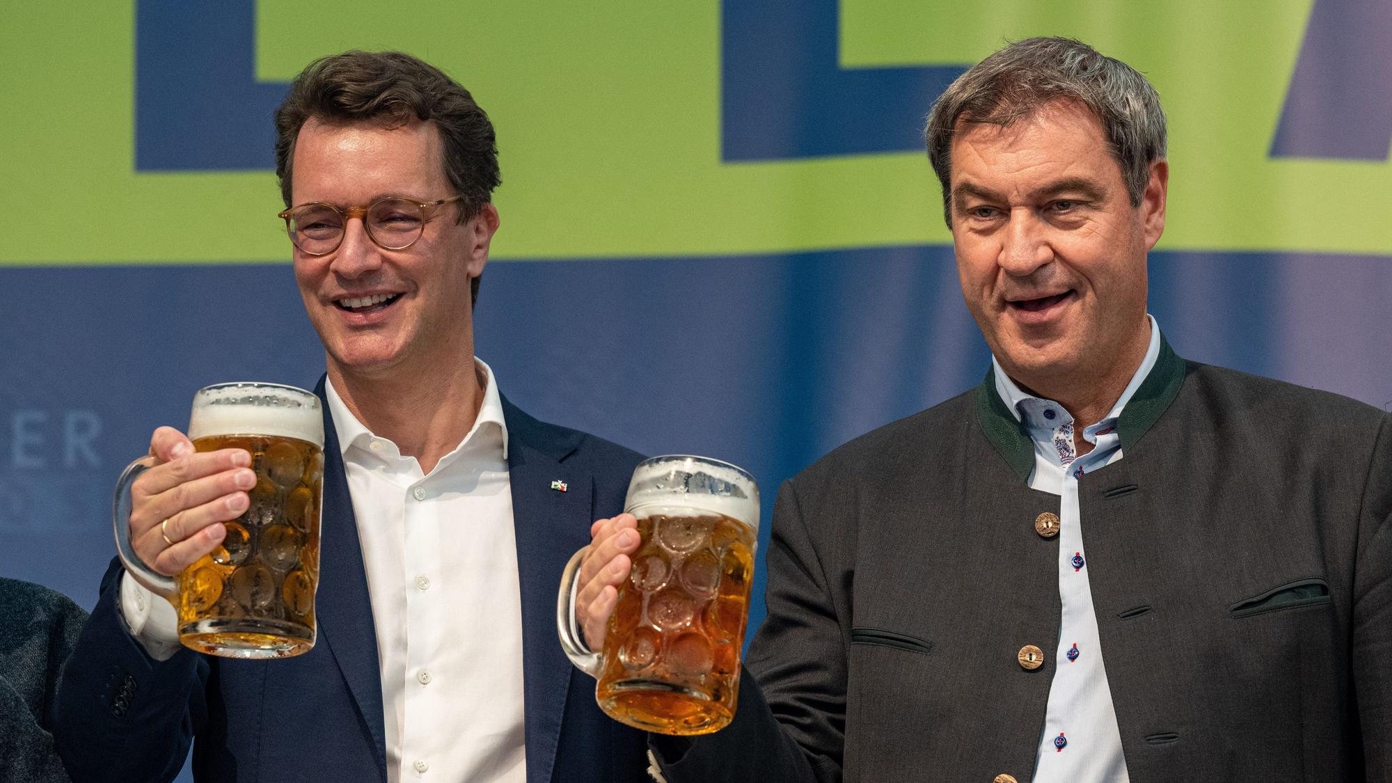 Markus Söder (r), Bayerns Ministerpräsident und CSU-Parteichef, und Hendrik Wüst, NRW-Ministerpräsident (CDU), stehen bei einem Volksfest mit Bierkrügen auf der Bühne.