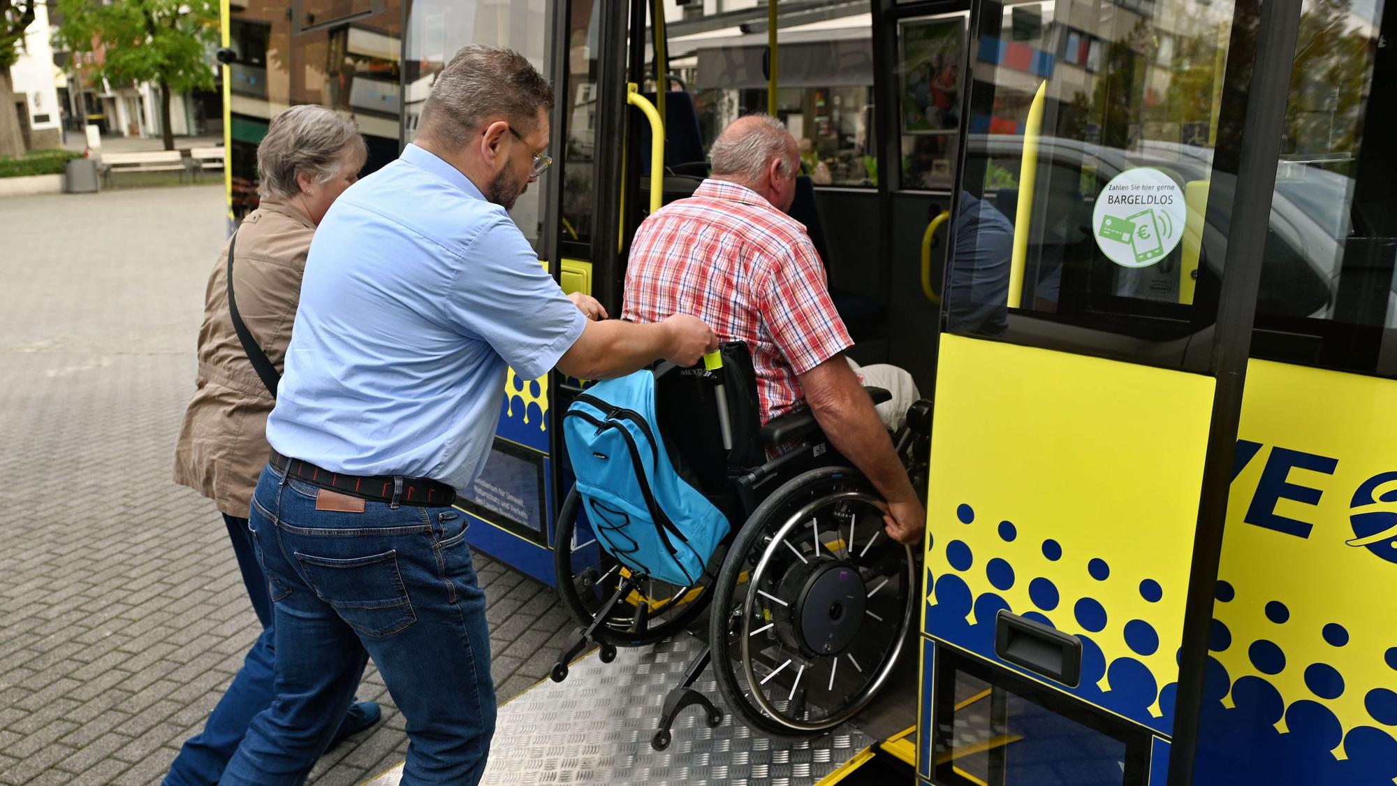 Zwei Leute schieben einen Rollstuhl über eine Rampe in den Bus.
