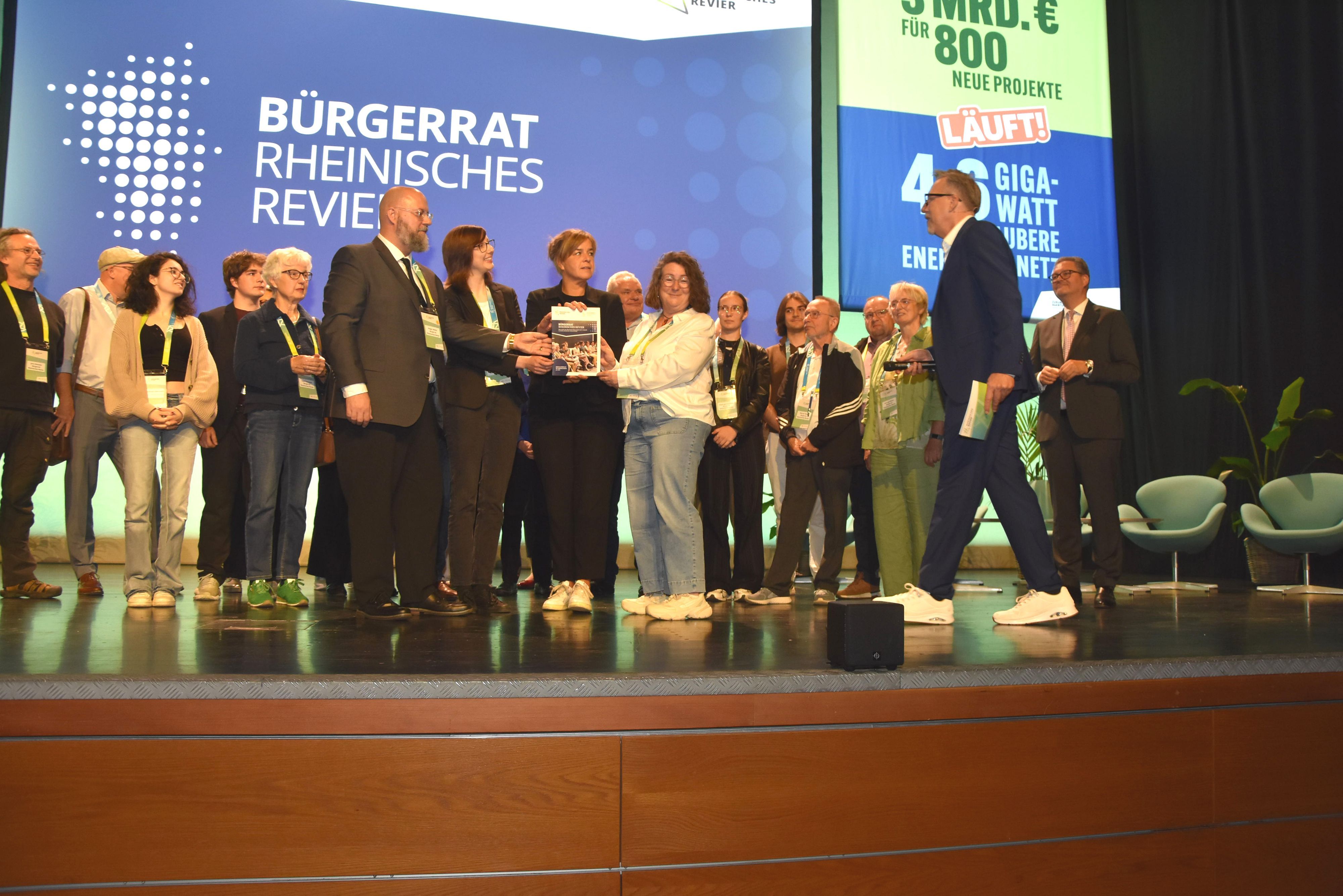 Das Bild zeigt den Bürgerrat auf der Bühne. Vertreter übergeben Ministerin Mona Neubaur einen Katalog mit 100 Vorschlägen für den Strukturwandel.