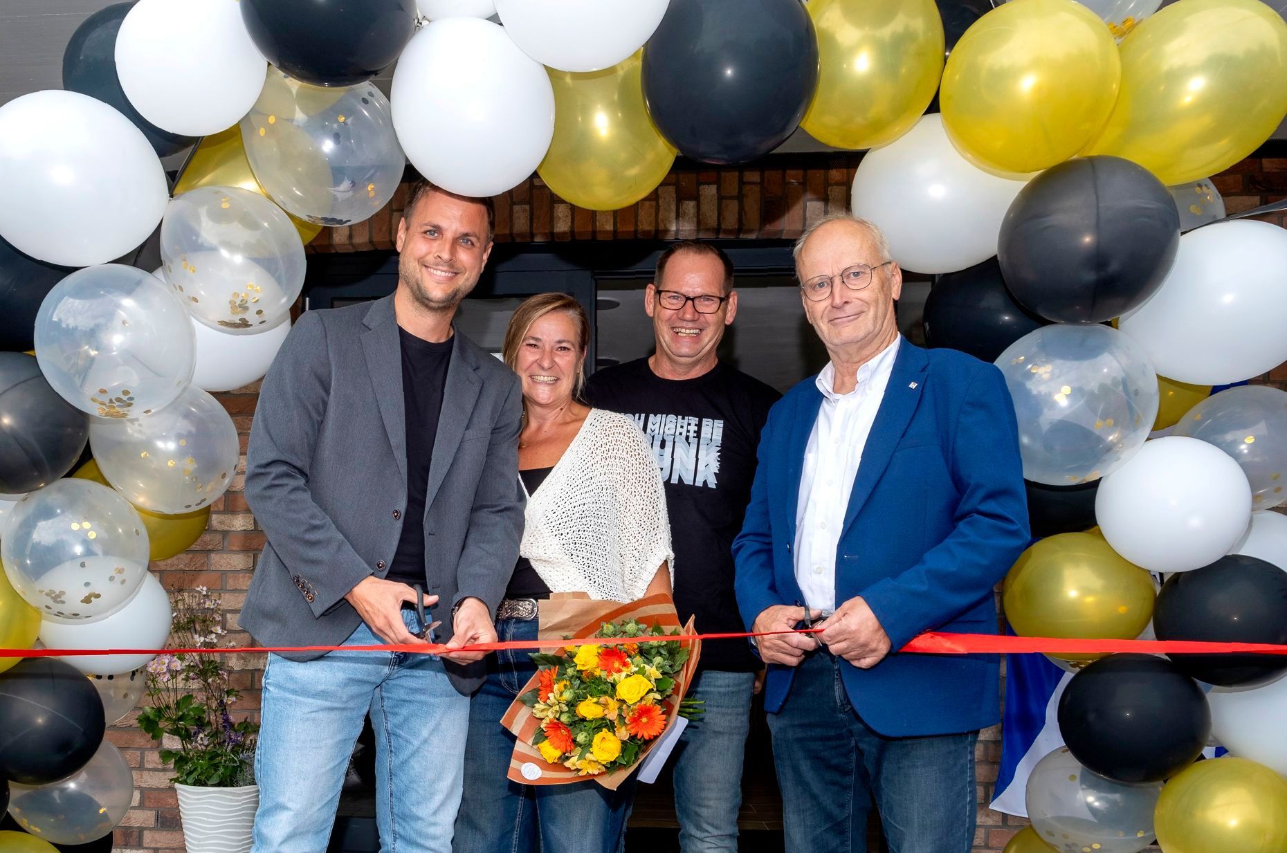 Die genannten Personen stehen vor einer mit Luftballons geschmückten Wand und durchschneiden gemeinsam ein rotes Band.
