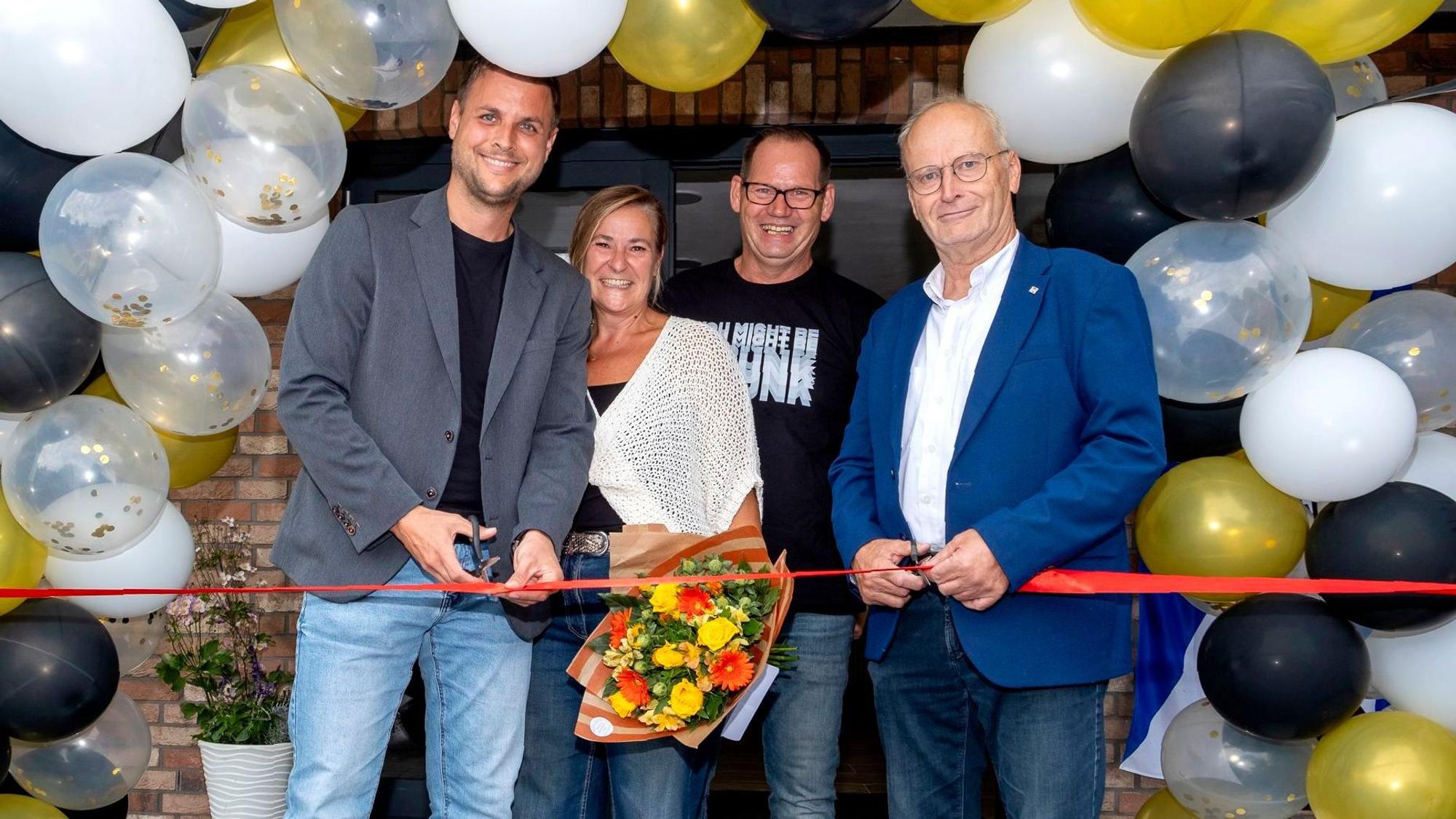 Die genannten Personen stehen vor einer mit Luftballons geschmückten Wand und durchschneiden gemeinsam ein rotes Band.