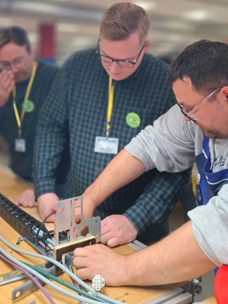 Ein Lebenshilfe-Mitarbeiter an einer Elektrokette erklärt zwei Currenta-Arbeitern die Arbeit.
