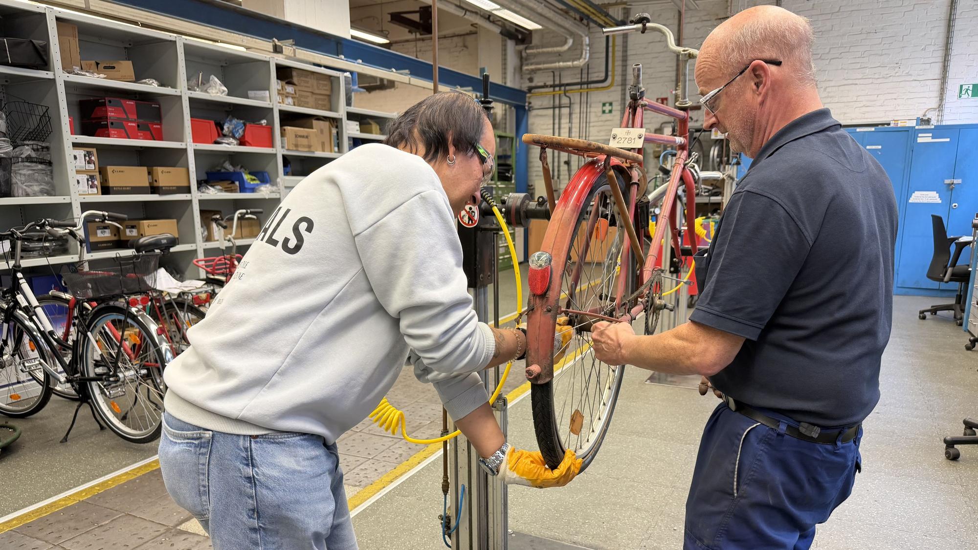Zwei Arbeiter in der Fahrradwerkstatt reparieren ein rotes Bayer-Rad.