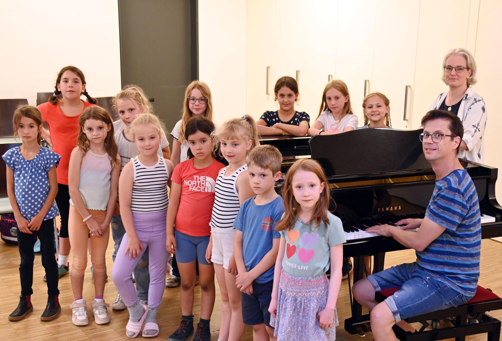 13 Kinder stehen um einen schwarzen Konzertflügel herum, an den Tasten sitzt der Leiter der Singschule Köln-Mitte Dirk-Johannes Neumann dahinter steht Stimmbildnerin Franzsika Schacht