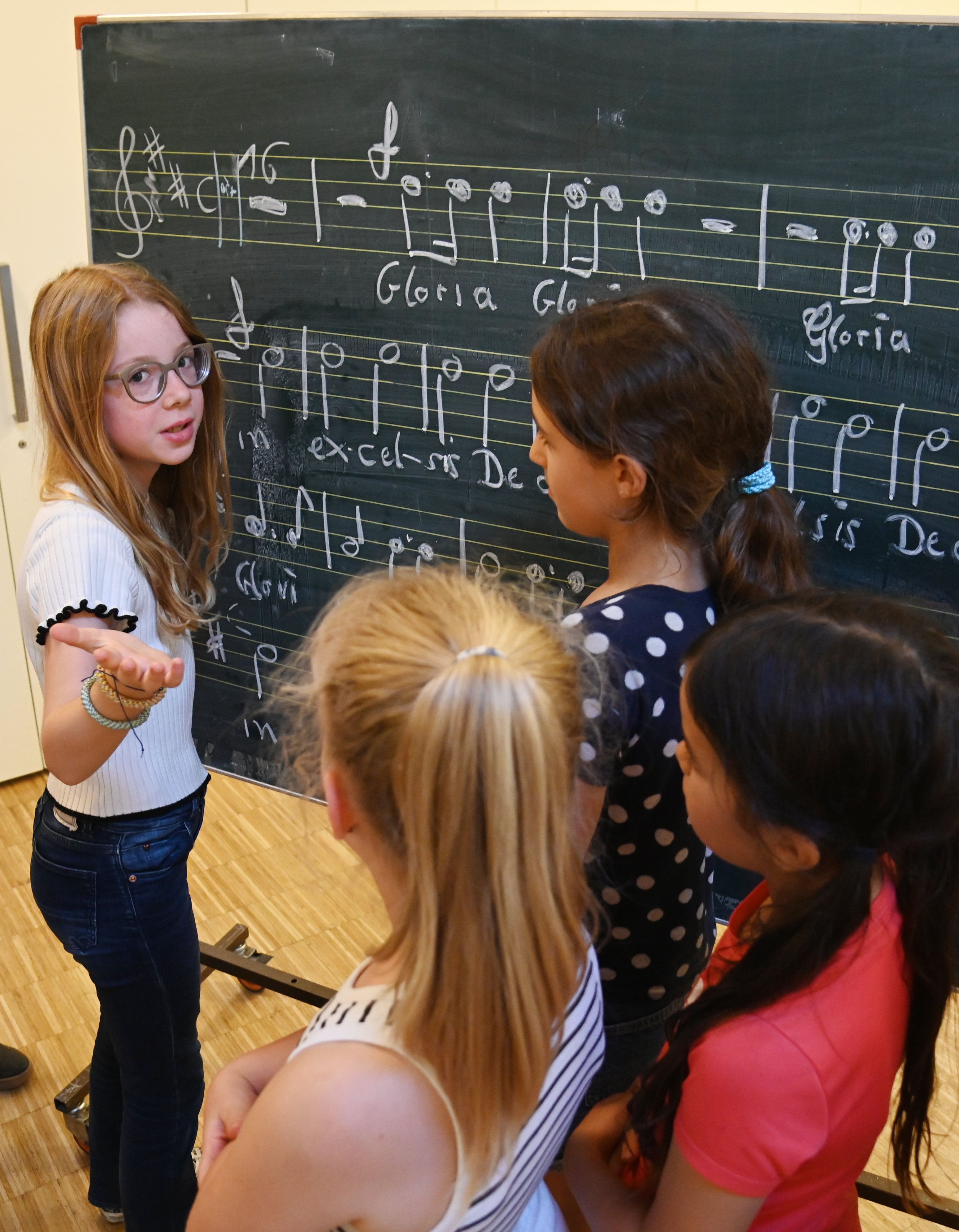 Vier Mädchen stehen vor einer Tafel, auf der die Noten des Lieds Gloria mit weißer Kreide aufgemalt sind - ein Kind erklärt, wie die Noten funktionieren.