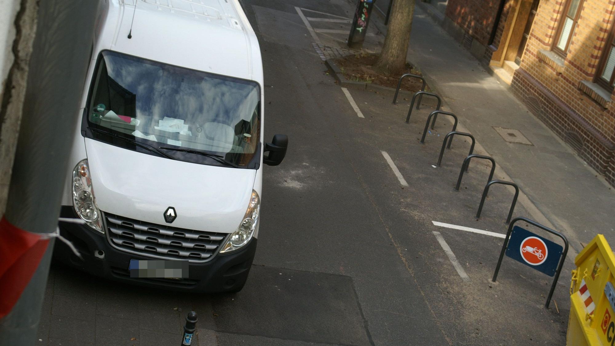 Ein weißer Transporter parkt in einer schmalen Straße gegenüber einem Fahrradparkplatz.