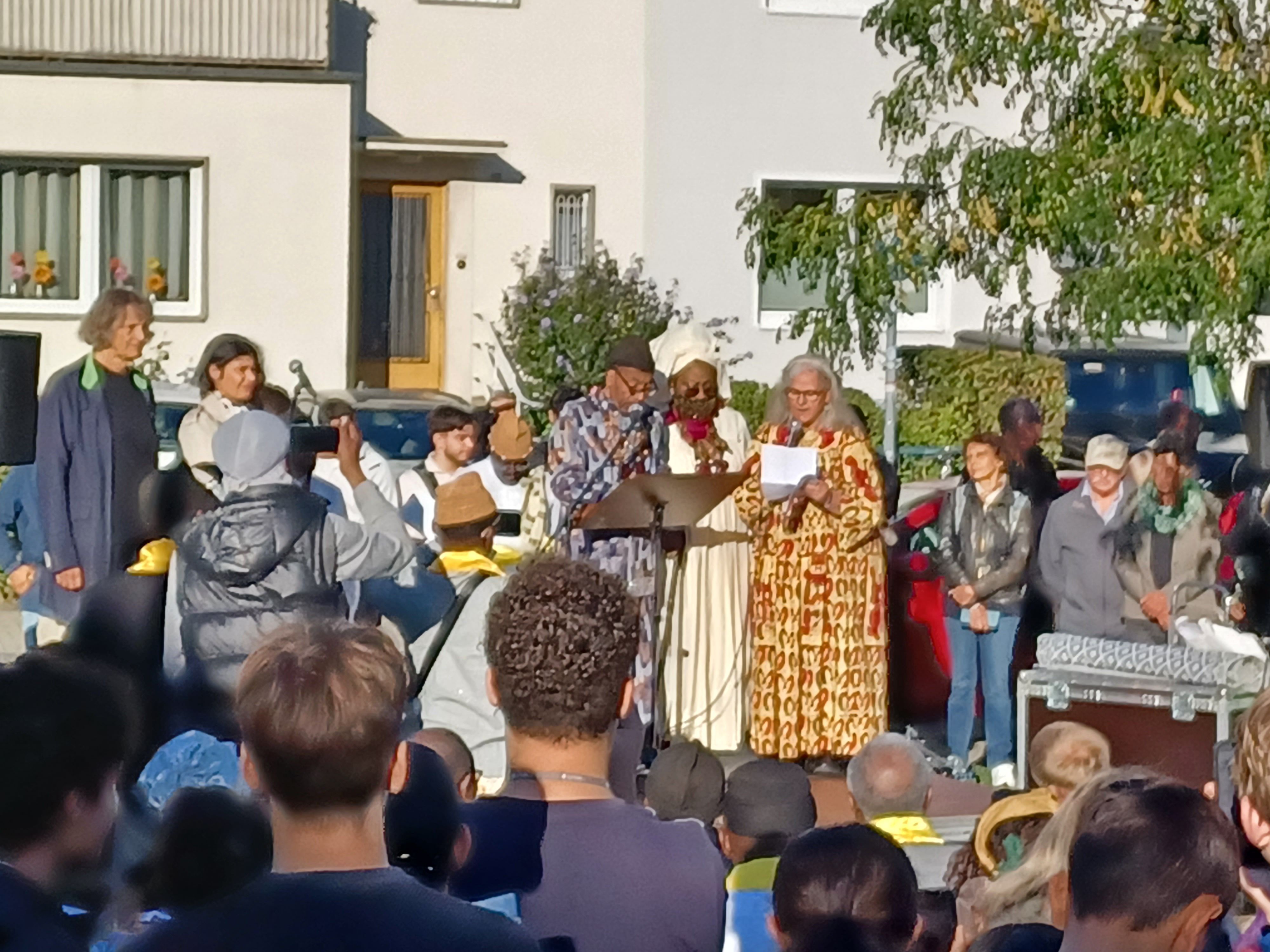 Viele Menschen stehen auf der Straße und hören drei in afrikanische Gewänder gekleideten Personen zu, die in Mikrofone sprechen.