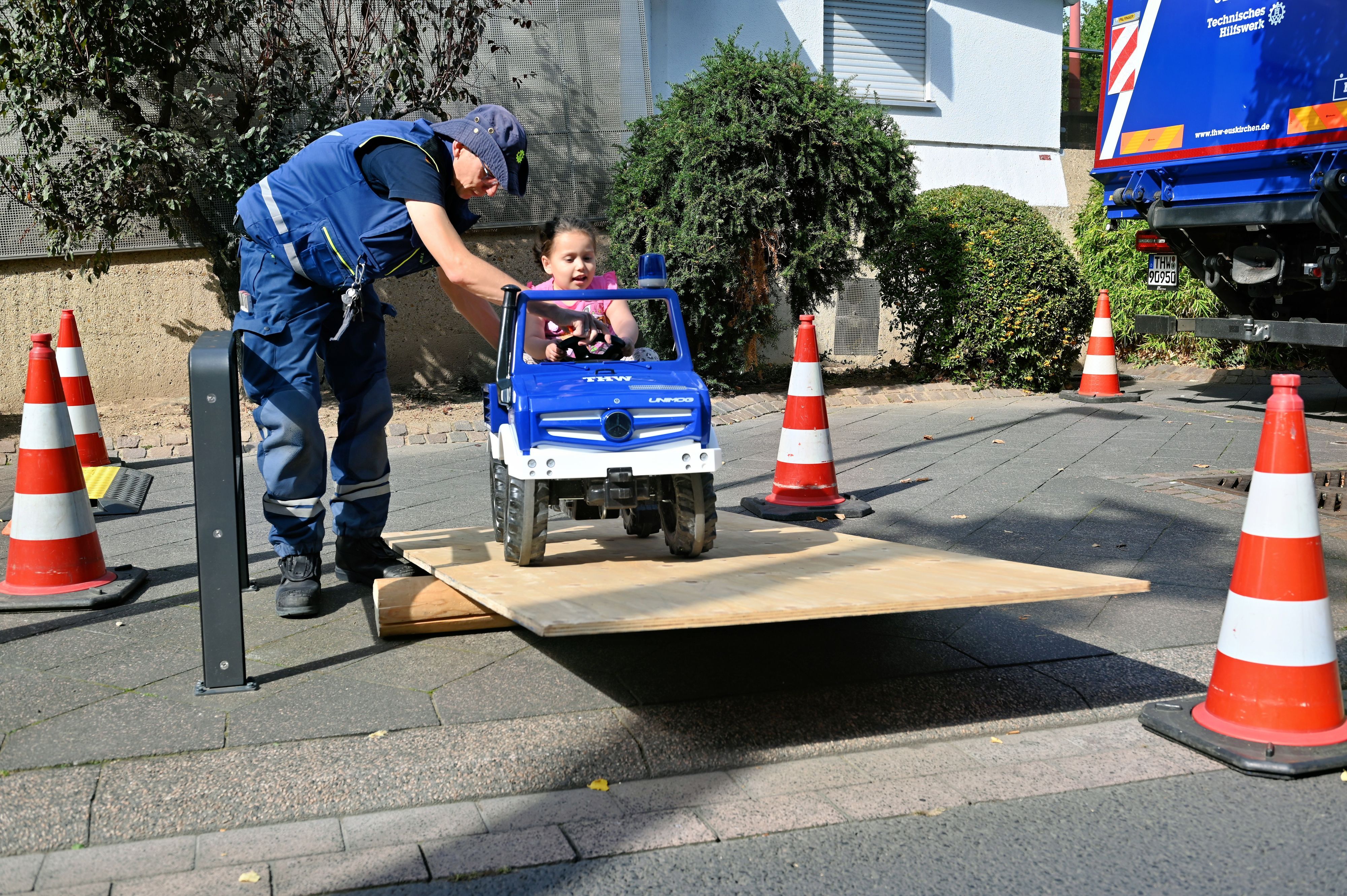 Ein THW-Helfer unterstützt ein Mädchen, das in einem Spielzeug-Lkw über ein Hindernis fährt.