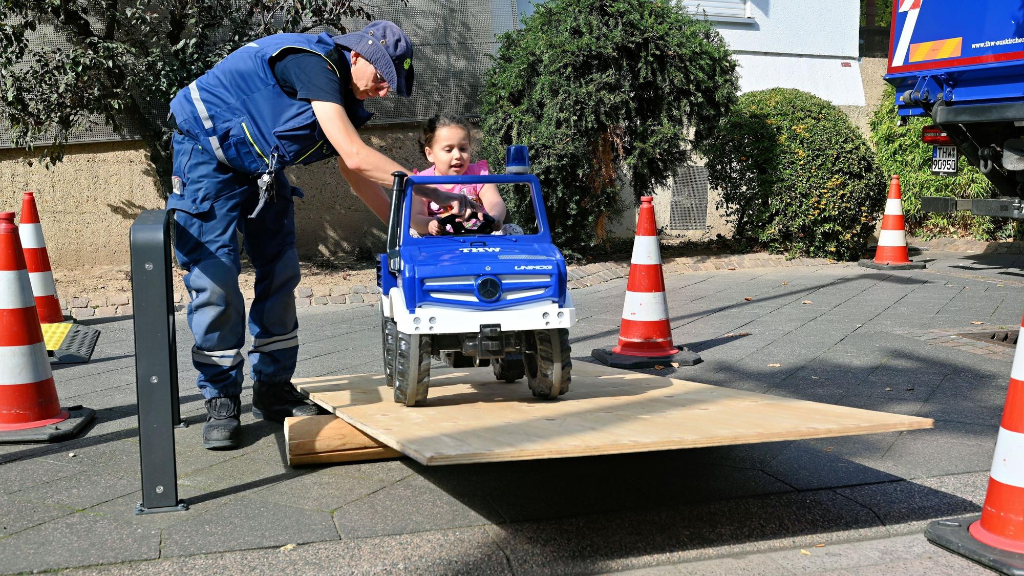Ein THW-Helfer unterstützt ein Mädchen, das in einem Spielzeug-Lkw über ein Hindernis fährt.