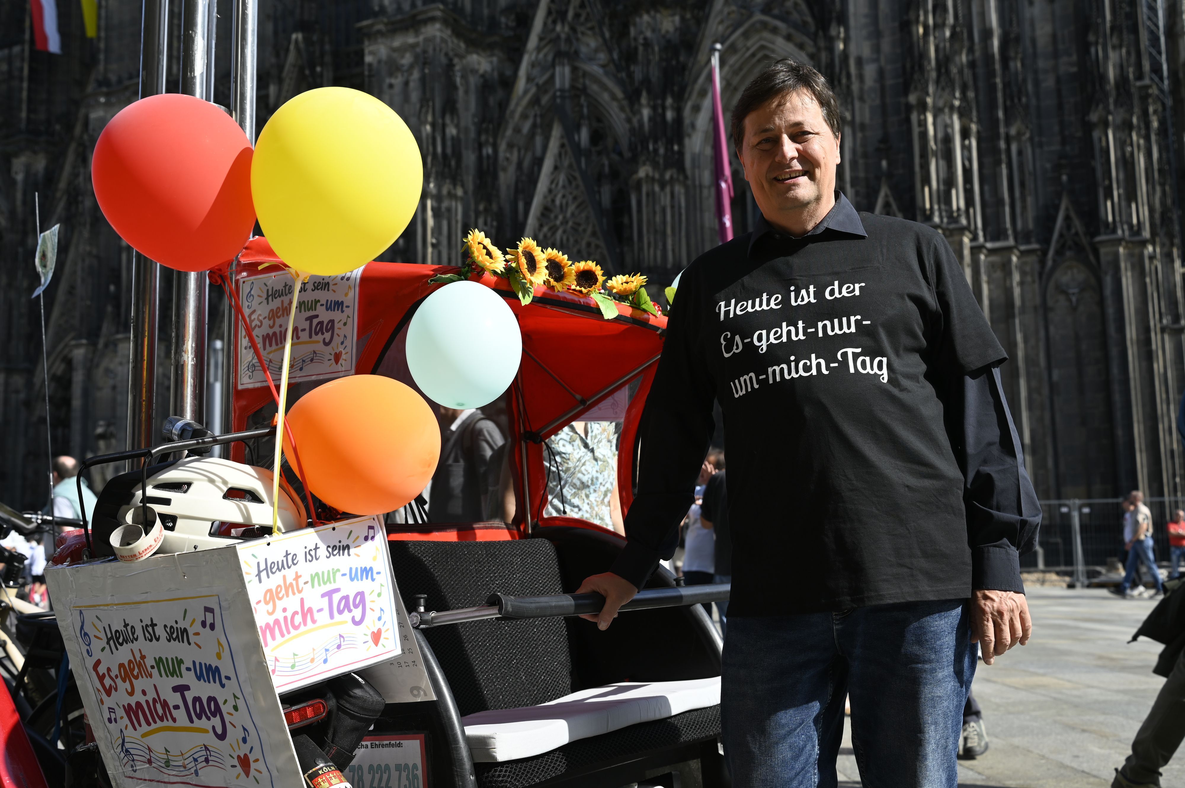 Ein Mann mit schwarzem Shirt und schwarzen Haaren steht neben einer geschmückten Rikscha vor dem Dom.