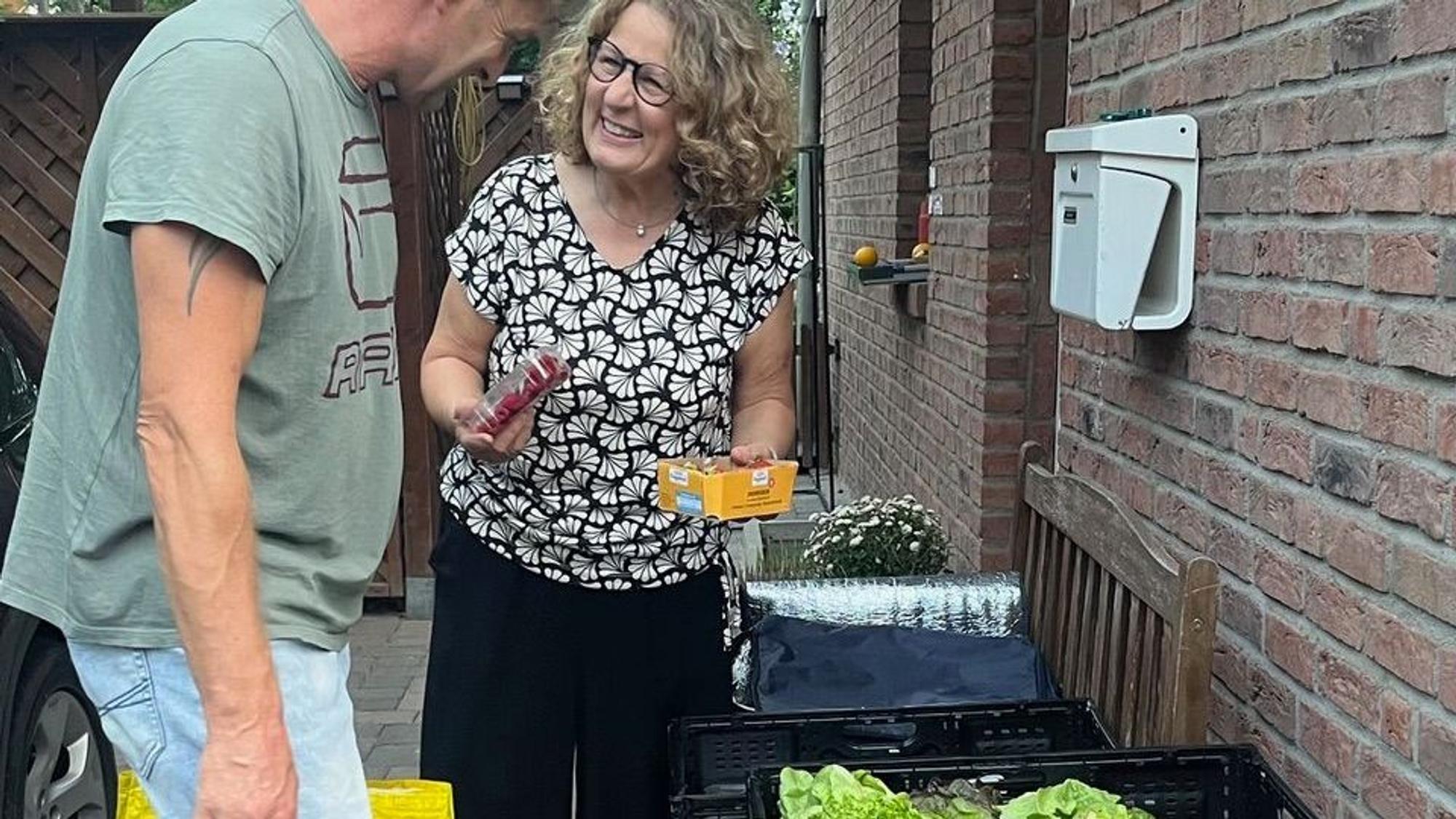 eine Frau reicht einem Mann einen Karton mit Pilzen. Beide stehen vor einer Hauswand