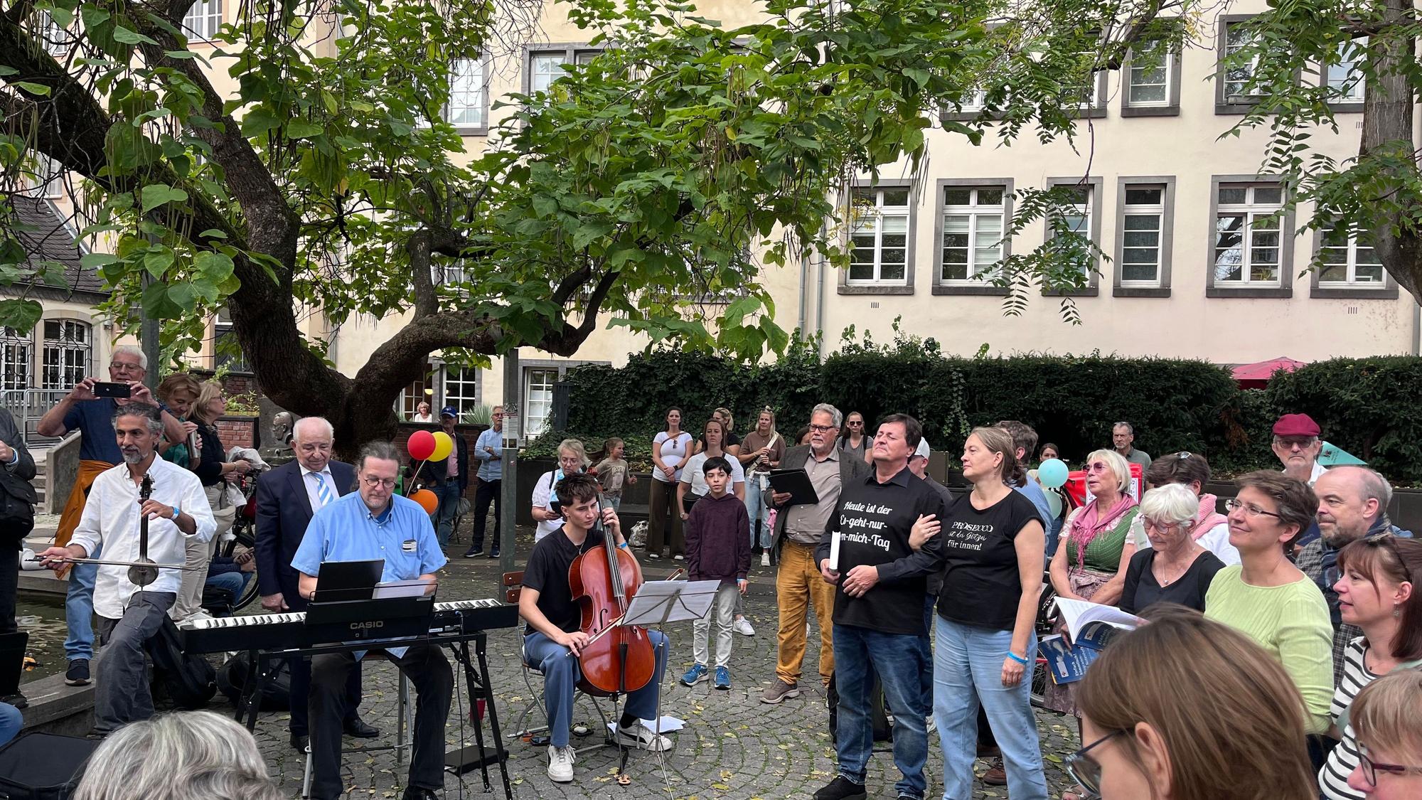 Viele Menschen stehen im Halbkreis. Unter einem alten Baum musizieren Musiker mit Keyboard, Cello und einem orientalischen Instrument.
